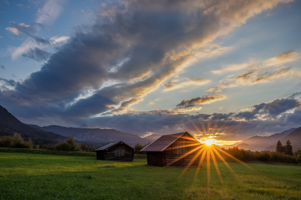 Sonnenuntergang im Werdenfelser Land bei Garmisch-Partenkirchen. Die tief stehende Sonne zaubert einen strahlenförmigen Sonnenstern zwischen traditionellen Holzhütten, grünen Wiesen und der Bergkulisse der bayerischen Alpen.