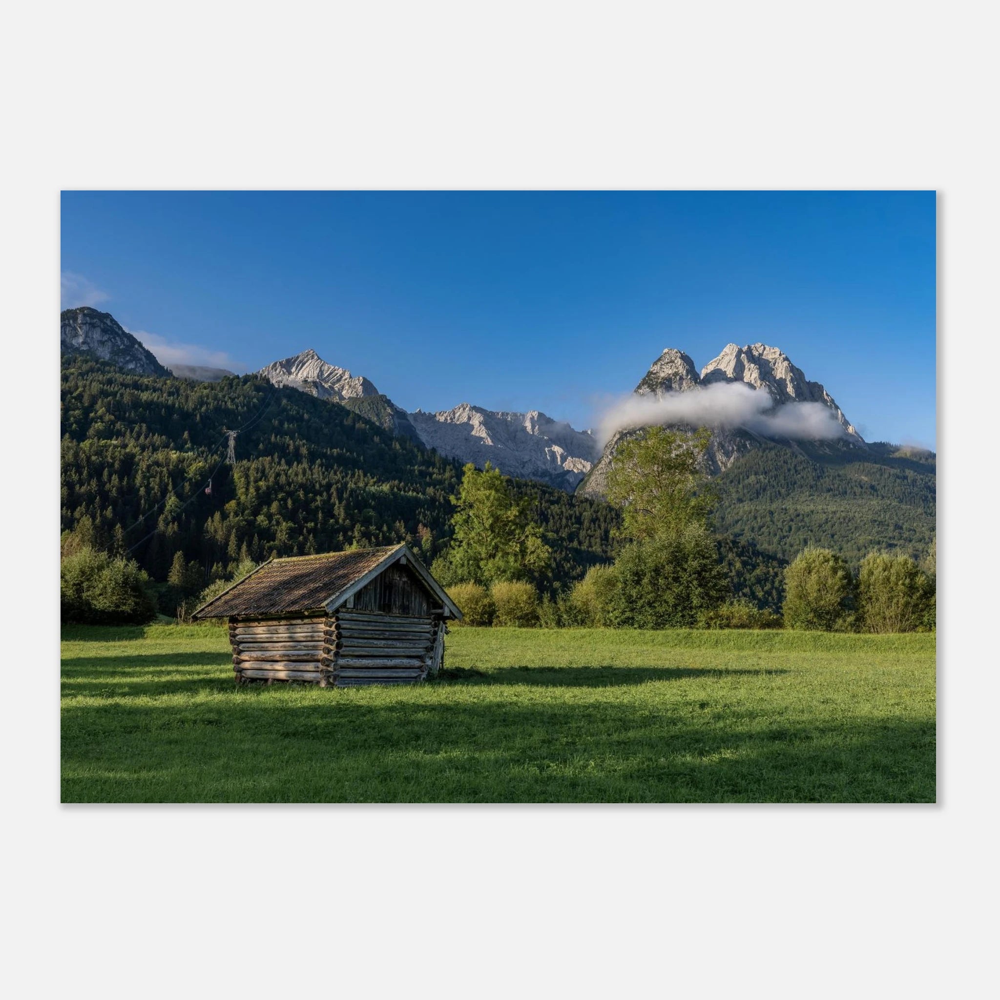 Panoramablick auf das Werdenfelser Land bei Garmisch-Partenkirchen mit einer kleinen Heuhütte auf einer grünen Wiese vor den Bergen des Wettersteingebirges. Warmes Morgenlicht, blauer Himmel und eine sanfte Wolke über den Gipfeln schaffen eine ruhige, natürliche Alpenstimmung.