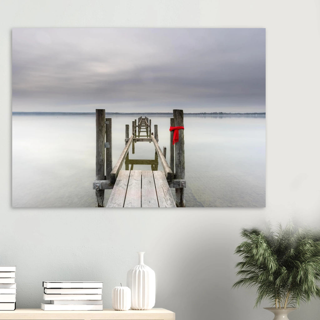 Alter Holzsteg am Ammersee mit einem leuchtend roten Schal, der an einem Pfosten befestigt ist. Die ruhige Langzeitbelichtung, das glatte Wasser und der sanft bewölkte Himmel schaffen eine stille, minimalistische Atmosphäre in Oberbayern.