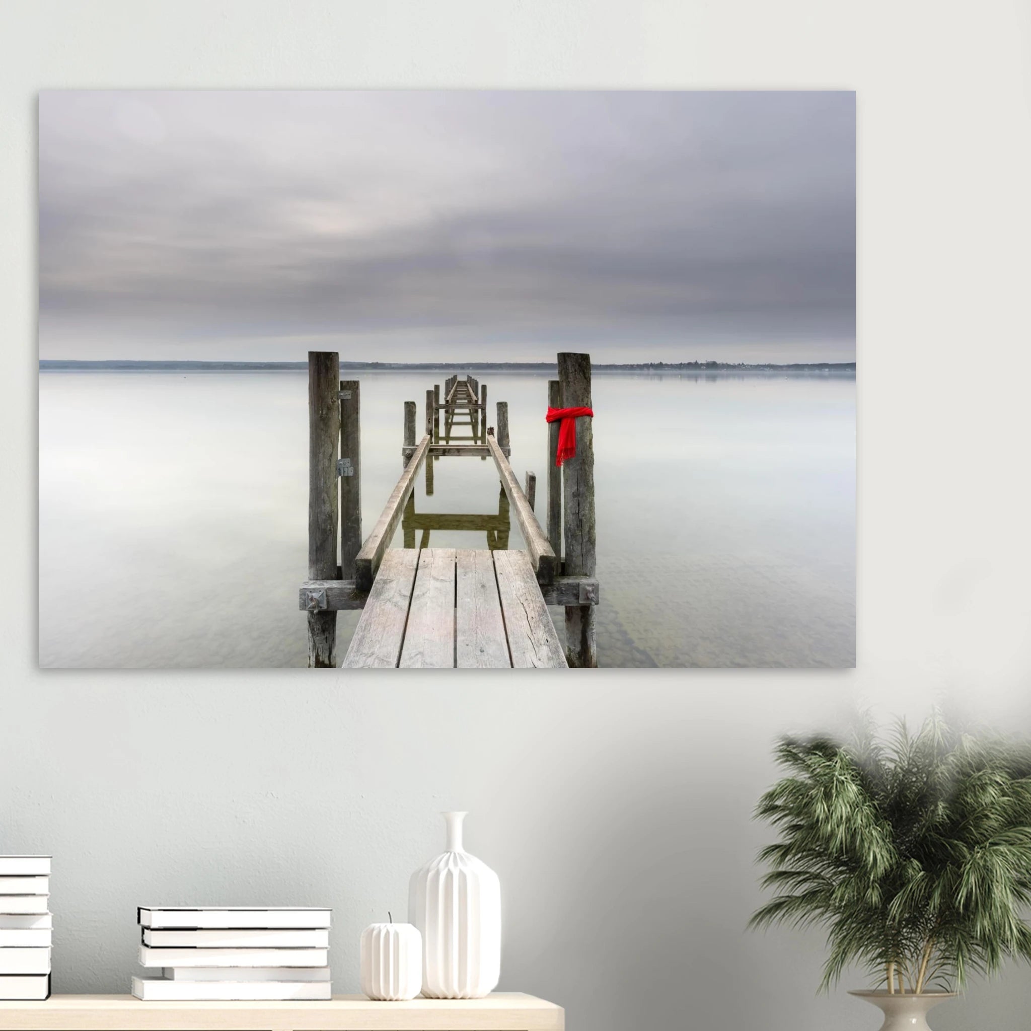 Alter Holzsteg am Ammersee mit einem leuchtend roten Schal, der an einem Pfosten befestigt ist. Die ruhige Langzeitbelichtung, das glatte Wasser und der sanft bewölkte Himmel schaffen eine stille, minimalistische Atmosphäre in Oberbayern.