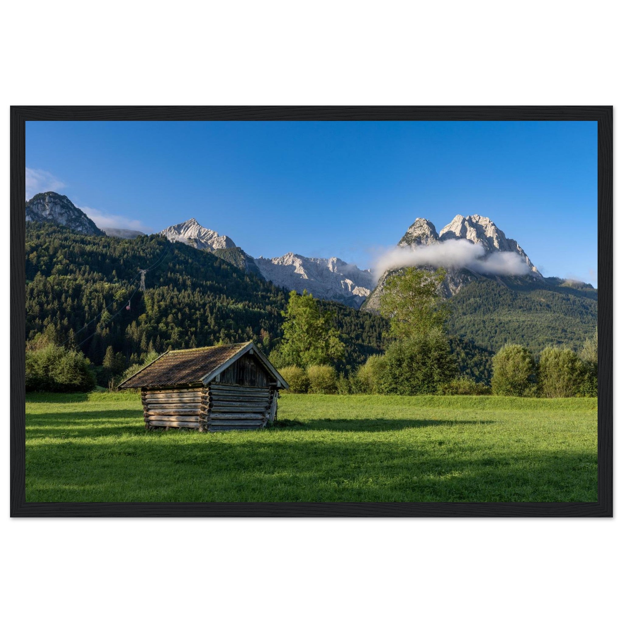 Gerahmtes Landschaftsposter  mit schwarzem Holzrahmen. Das Motiv zeigt eine kleine Holzhütte auf einer grünen Wiese vor einer imposanten Bergkulisse in Garmisch-Partenkirchen, unter tiefblauem Himmel und sanften Wolken – eine natürliche Szene voller Ruhe, Weite und alpiner Atmosphäre.