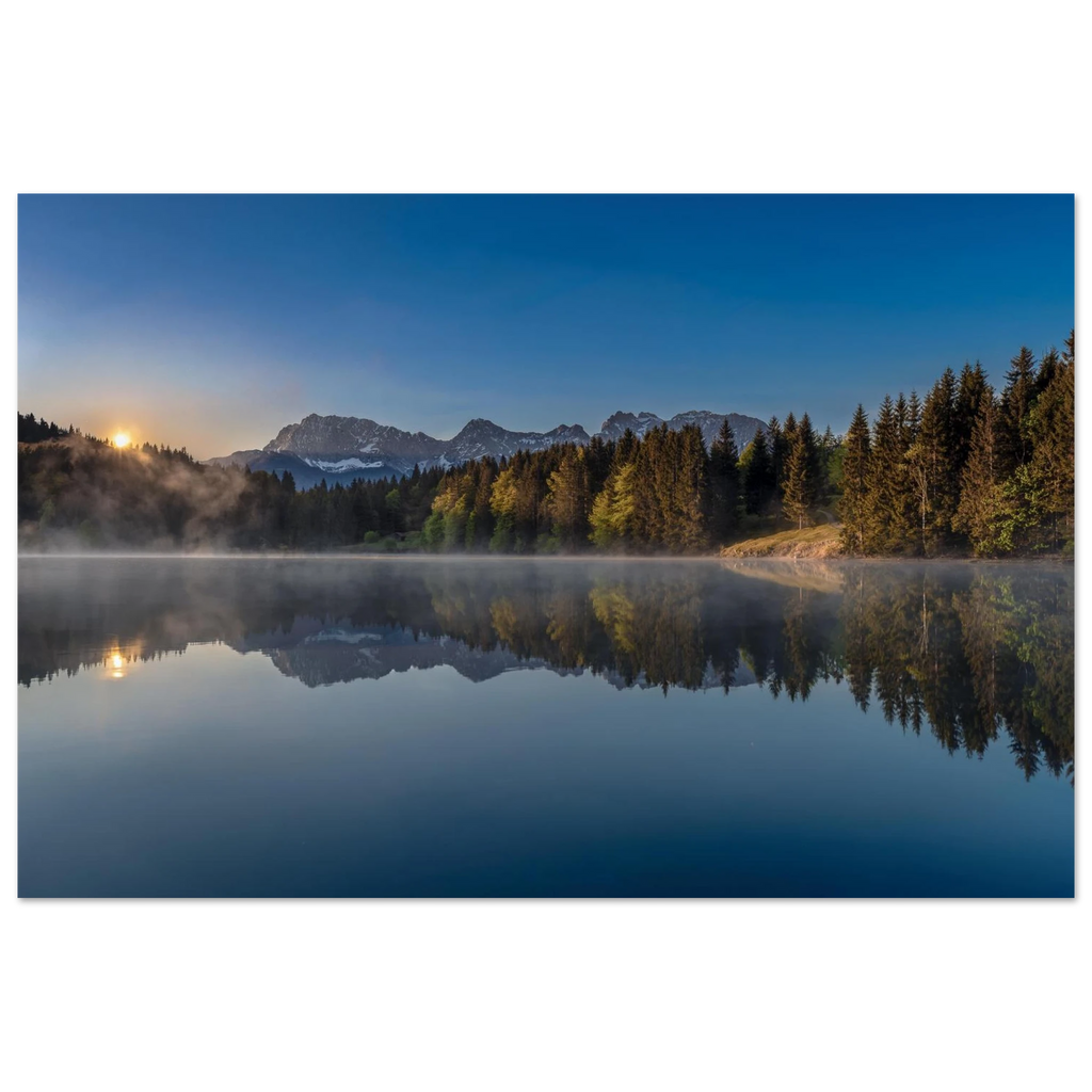 Ein stiller Morgen am Geroldsee in Oberbayern. Feine Nebelschleier ziehen sanft über das ruhige Wasser, während sich die umliegenden Wälder und die Bergwelt nahezu vollkommen im See spiegeln. Das erste warme Licht des Tages berührt die Bäume und schenkt der Landschaft eine besondere Ruhe und Tiefe.
