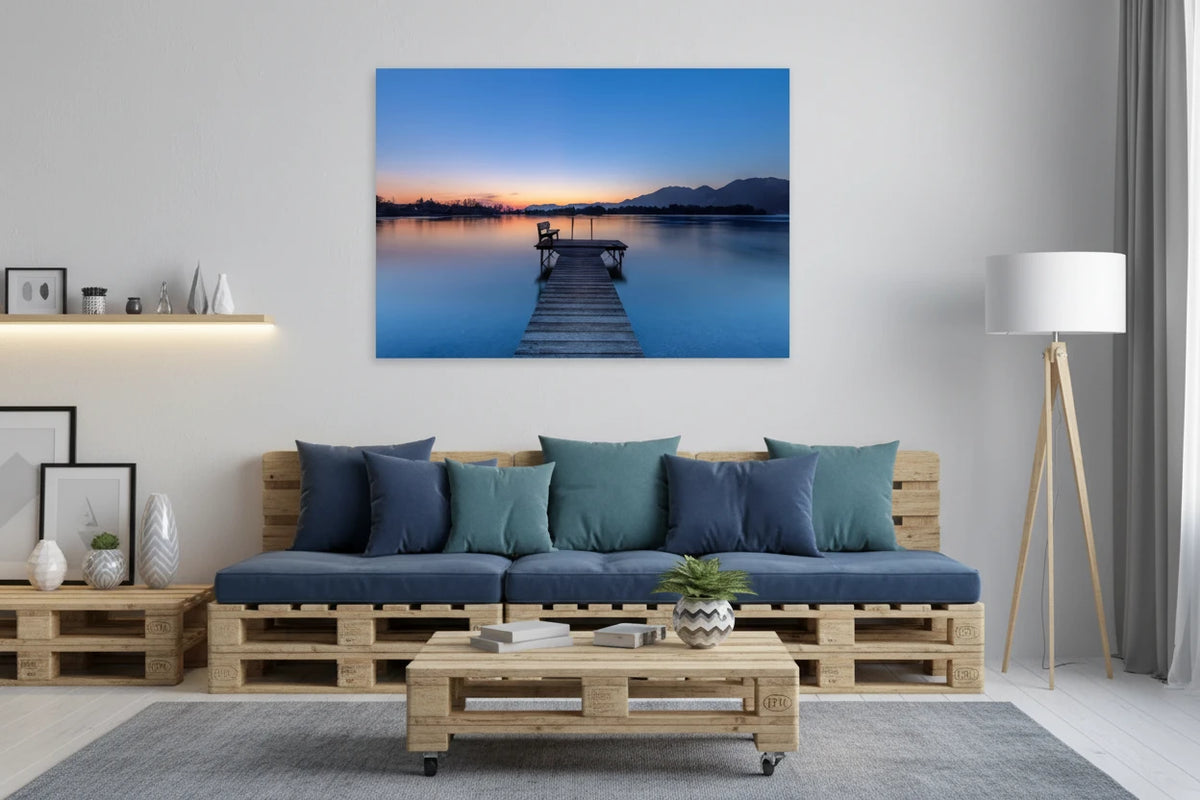 Aluminium-Print mit Sonnenaufgang am Kochelsee: Ruhiger See in sanften Blau- und Orangetönen, ein hölzerner Steg mit Bank führt ins stille Wasser, im Hintergrund Berge und die zarte Morgenstimmung des beginnenden Tages.