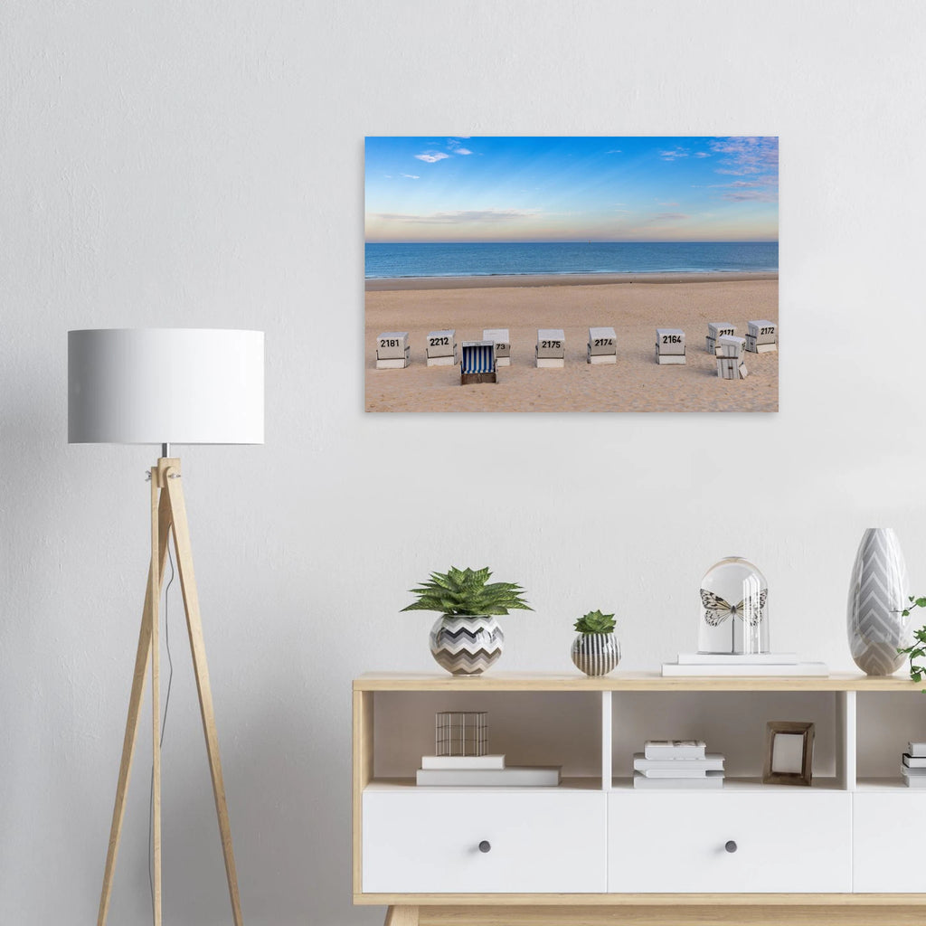 Das Motiv zeigt einen weiten, ruhigen Strand in Westerland auf Sylt in einer klaren, sommerlichen Stimmung. Im Vordergrund liegt der helle, fein strukturierte Sand, in dem mehrere weiße Strandkörbe in einer lockeren Reihe stehen. Einer davon ist geöffnet und mit blau-weiß gestreiftem Innenstoff ein besonderer Blickfang.
