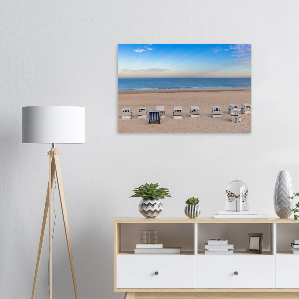 Das Motiv zeigt einen weiten, ruhigen Strand in Westerland auf Sylt in einer klaren, sommerlichen Stimmung. Im Vordergrund liegt der helle, fein strukturierte Sand, in dem mehrere weiße Strandkörbe in einer lockeren Reihe stehen. Einer davon ist geöffnet und mit blau-weiß gestreiftem Innenstoff ein besonderer Blickfang.
