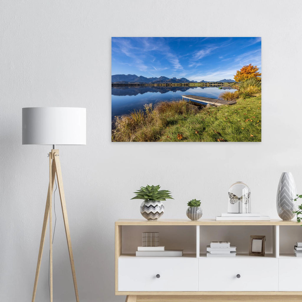Herbstlicher Hopfensee mit ruhigem, spiegelndem Wasser, Holzsteg und leuchtend goldenem Baum vor der klaren Kulisse der Allgäuer Alpen.

