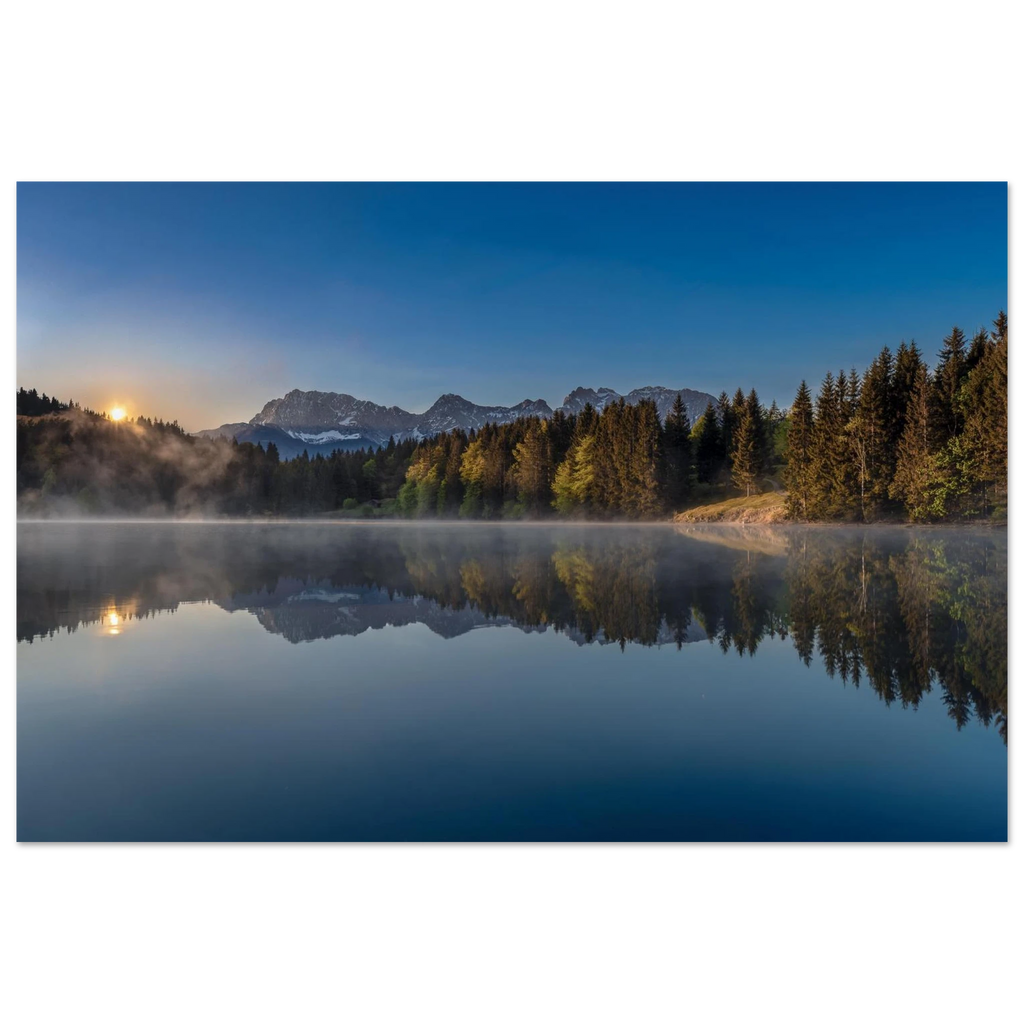 Ein stiller Morgen am Geroldsee in Oberbayern. Feine Nebelschleier ziehen sanft über das ruhige Wasser, während sich die umliegenden Wälder und die Bergwelt nahezu vollkommen im See spiegeln. Das erste warme Licht des Tages berührt die Bäume und schenkt der Landschaft eine besondere Ruhe und Tiefe.
