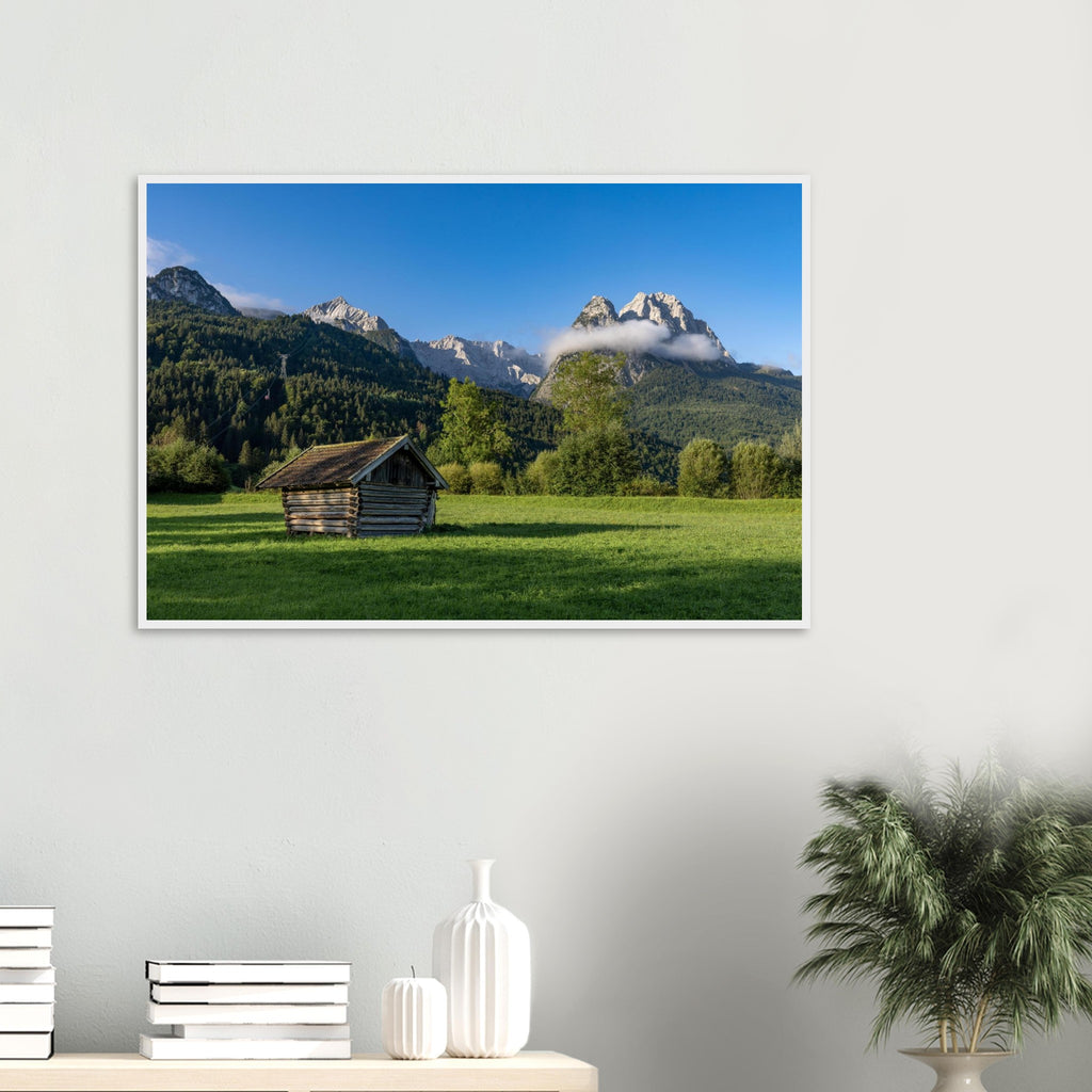 Gerahmtes Landschaftsposter im Format 60x90 cm mit weissem Holzrahmen. Das Motiv zeigt eine kleine Holzhütte auf einer grünen Wiese vor einer imposanten Bergkulisse in Garmisch-Partenkirchen, unter tiefblauem Himmel und sanften Wolken – eine natürliche Szene voller Ruhe, Weite und alpiner Atmosphäre.