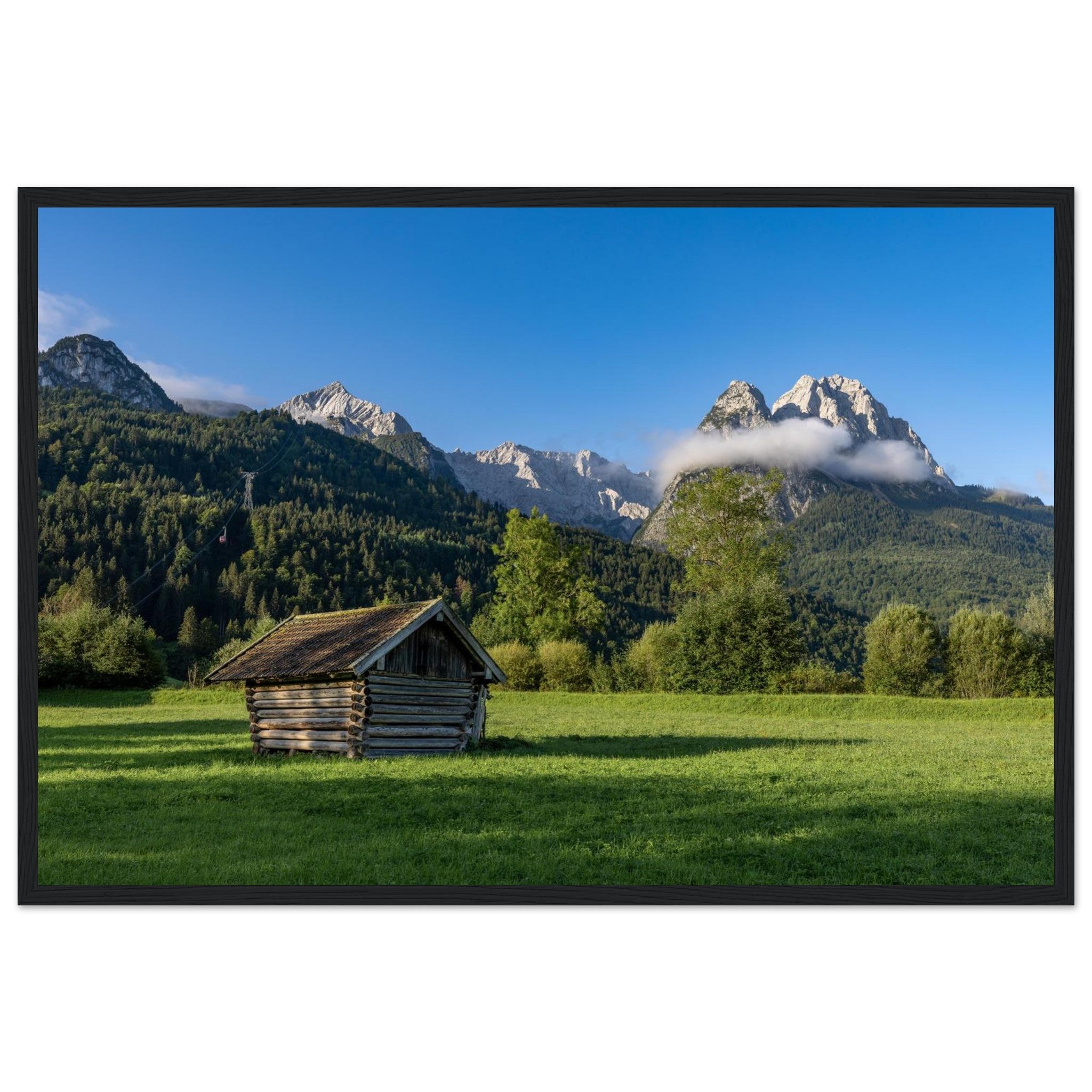 Gerahmtes Landschaftsposter mit schwarzem Holzrahmen im Format 60x90 cm . Das Motiv zeigt eine kleine Holzhütte auf einer Wiese vor beeindruckender Bergkulisse unter blauem Himmel – eingebettet in natürliche Materialien und eine ruhige, harmonische Wohnatmosphäre.