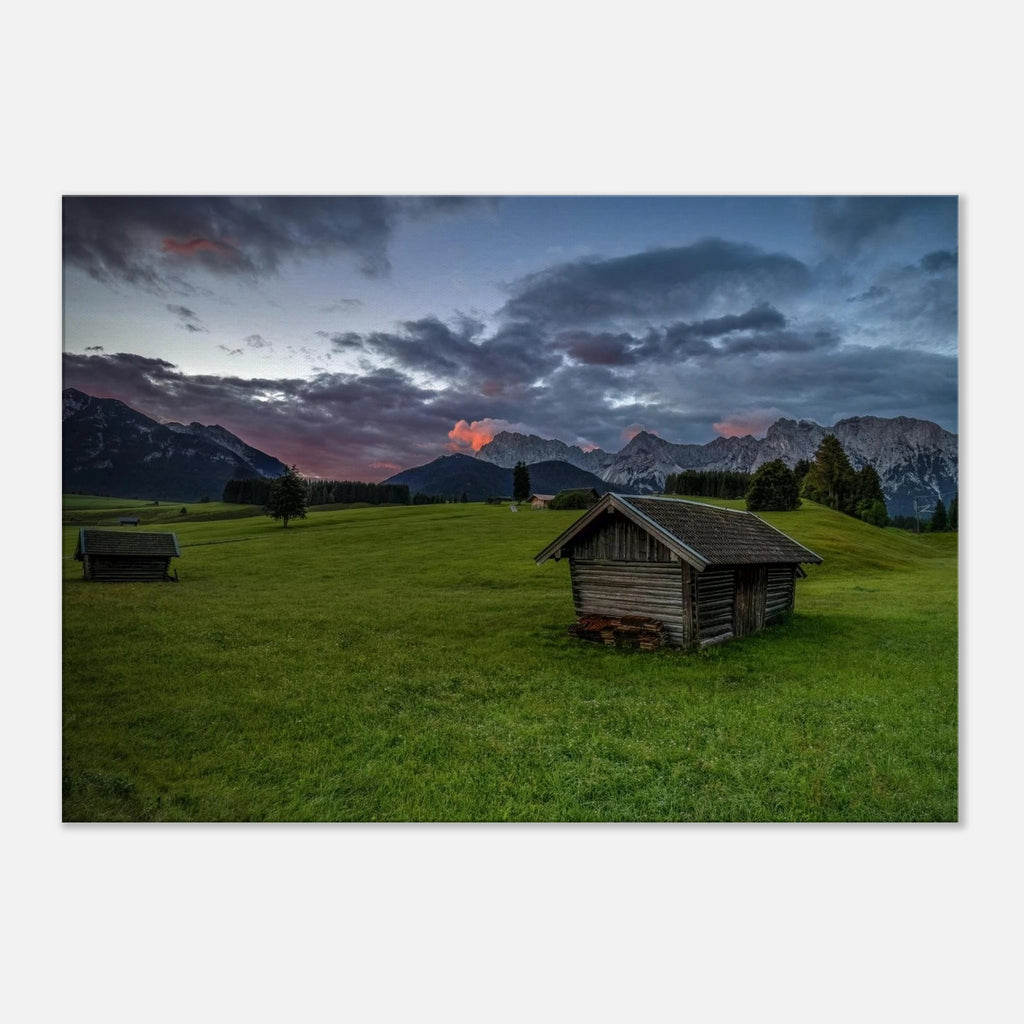 Grüne Buckelwiesen bei Krün mit traditionellen Heustadeln, dem Karwendelgebirge im Hintergrund und dramatischen Regenwolken in der frühen Morgenstimmung.
