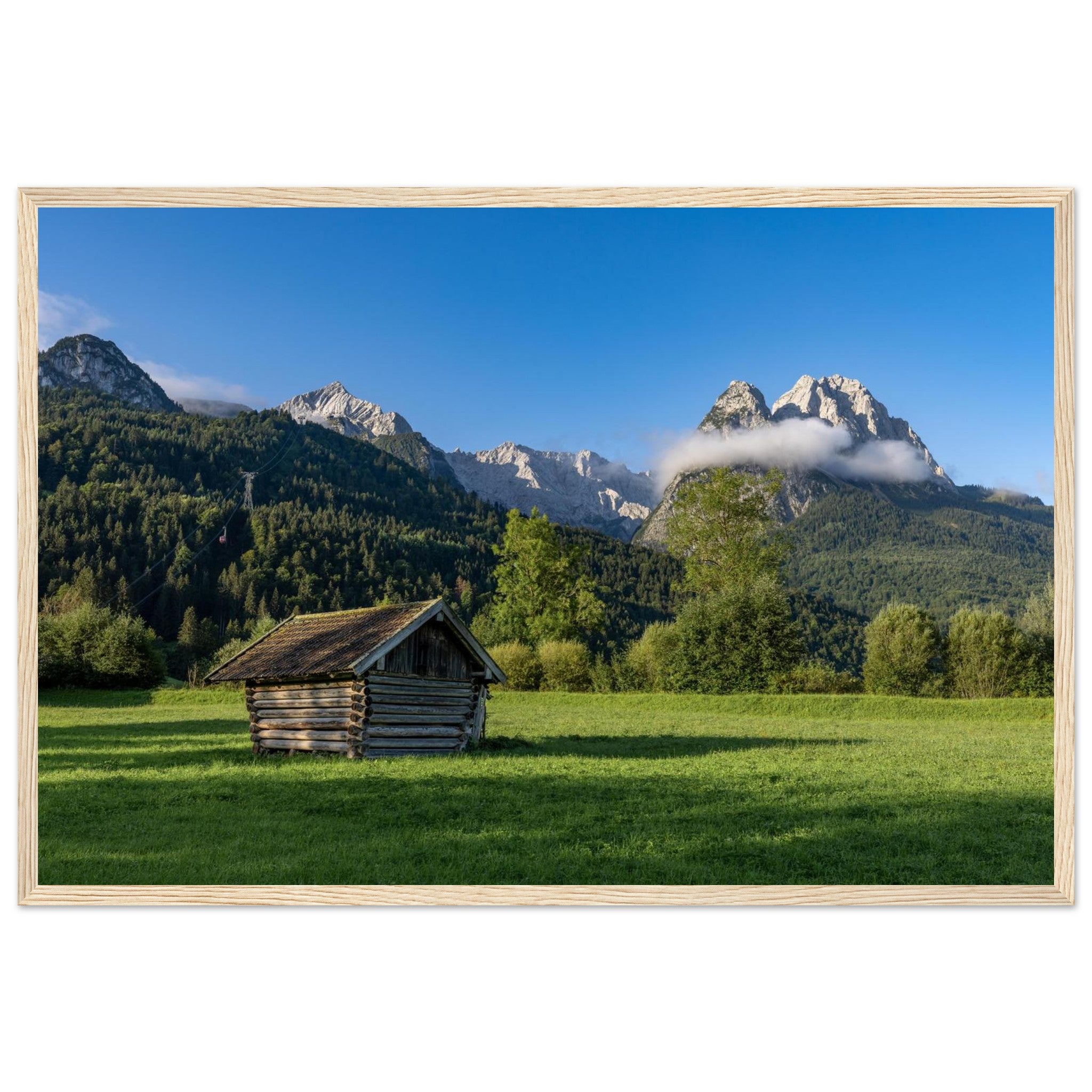Gerahmtes Landschaftsposter mit naturfarbenen Holzrahmen im Format 60x90 cm Das Motiv zeigt eine kleine Holzhütte auf einer Wiese vor beeindruckender Bergkulisse unter blauem Himmel – eingebettet in natürliche Materialien und eine ruhige, harmonische Wohnatmosphäre.