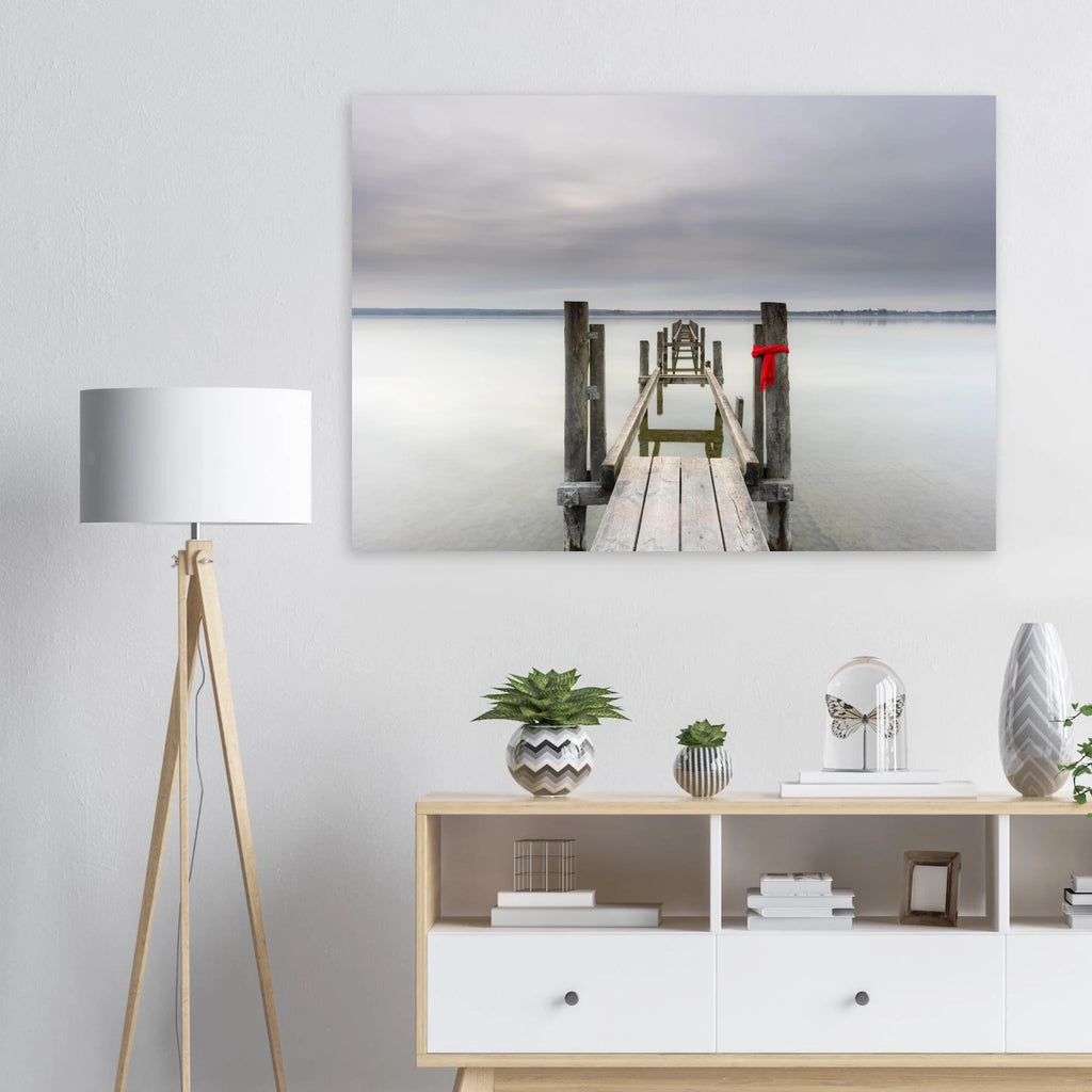 Alter Holzsteg am Ammersee mit einem leuchtend roten Schal, der an einem Pfosten befestigt ist. Die ruhige Langzeitbelichtung, das glatte Wasser und der sanft bewölkte Himmel schaffen eine stille, minimalistische Atmosphäre in Oberbayern.