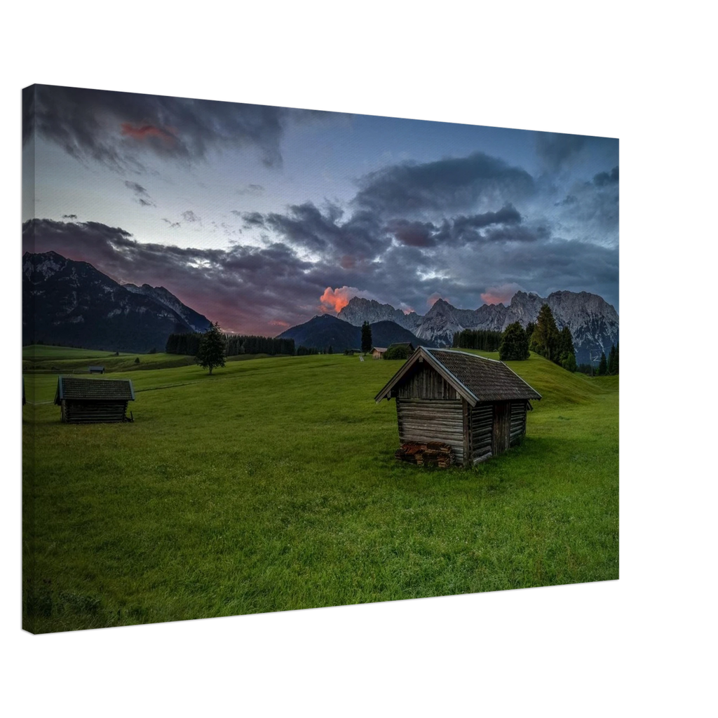 Grüne Buckelwiesen bei Krün mit traditionellen Heustadeln, dem Karwendelgebirge im Hintergrund und dramatischen Regenwolken in der frühen Morgenstimmung.
