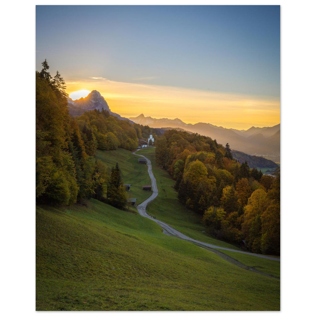 Vertikales Landschaftsposter mit einem stimmungsvollen Sonnenuntergang im Werdenfelserland. Ein geschwungener Weg führt durch Wiesen und herbstlich gefärbte Wälder hinauf nach Wamberg, dem höchstgelegenen Kirchdorf Deutschlands. Die kleine Kirche liegt eingebettet in die Berglandschaft, während die untergehende Sonne den Himmel in warme Gold- und Orangetöne taucht.