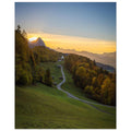 Vertikales Landschaftsposter mit einem stimmungsvollen Sonnenuntergang im Werdenfelserland. Ein geschwungener Weg führt durch Wiesen und herbstlich gefärbte Wälder hinauf nach Wamberg, dem höchstgelegenen Kirchdorf Deutschlands. Die kleine Kirche liegt eingebettet in die Berglandschaft, während die untergehende Sonne den Himmel in warme Gold- und Orangetöne taucht.
