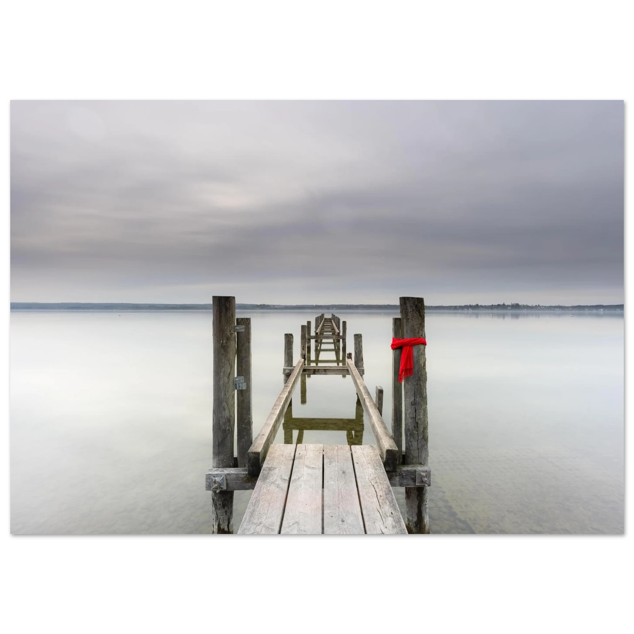 Alter Holzsteg am Ammersee mit einem leuchtend roten Schal, der an einem Pfosten befestigt ist. Die ruhige Langzeitbelichtung, das glatte Wasser und der sanft bewölkte Himmel schaffen eine stille, minimalistische Atmosphäre in Oberbayern.