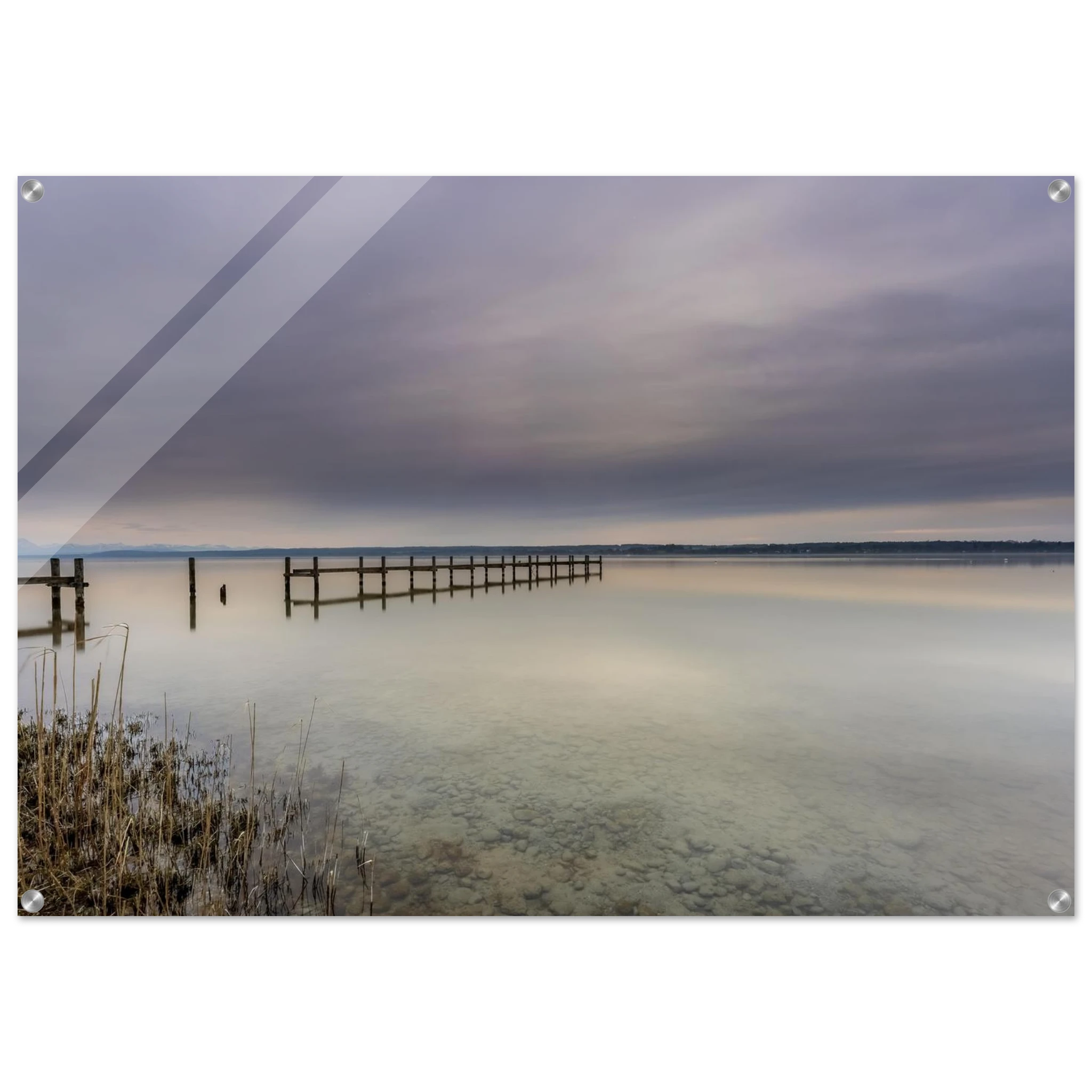 Ruhige Stimmung am Ammersee mit einem langen Holzsteg, der weit in das spiegelglatte Wasser hinausführt. Im Vordergrund sind Schilf und die klar sichtbaren Steine des flachen Uferbereichs zu erkennen. Über dem See liegt ein sanft bewölkter Himmel in zarten Grau-, Blau- und Beigetönen, dessen Licht sich im Wasser spiegelt. Die weite, reduzierte Landschaft vermittelt Stille, Gelassenheit und die zeitlose Schönheit eines friedlichen Moments am Ammersee.