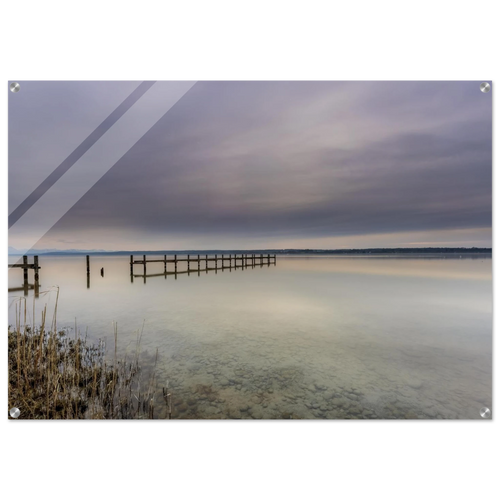Ruhige Stimmung am Ammersee mit einem langen Holzsteg, der weit in das spiegelglatte Wasser hinausführt. Im Vordergrund sind Schilf und die klar sichtbaren Steine des flachen Uferbereichs zu erkennen. Über dem See liegt ein sanft bewölkter Himmel in zarten Grau-, Blau- und Beigetönen, dessen Licht sich im Wasser spiegelt. Die weite, reduzierte Landschaft vermittelt Stille, Gelassenheit und die zeitlose Schönheit eines friedlichen Moments am Ammersee.