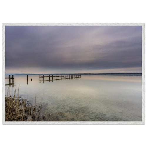 Ammersee Premium Matt Poster mit weißem Holzrahmen. Die Fotografie zeigt einen ruhigen Steg, der weit in das spiegelglatte Wasser des Ammersees hinausführt. Zarte Grau-, Blau- und Cremetöne prägen den bewölkten Himmel und spiegeln sich sanft auf der Wasseroberfläche. Im Vordergrund sind Schilf und die klaren Kieselsteine unter dem flachen Wasser sichtbar. Die minimalistische Landschaft vermittelt Stille, Weite und eine entspannte Atmosphäre und wirkt als elegante Wanddekoration in natürlichen Wohnräumen.