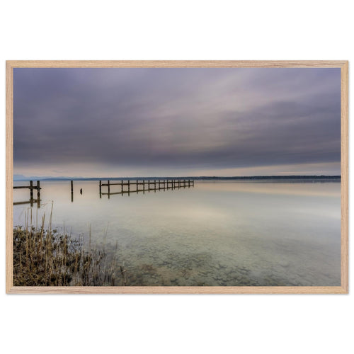 Ammersee Premium Matt Poster mit naturfarbeHolzrahmen. Die Fotografie zeigt einen ruhigen Steg, der weit in das spiegelglatte Wasser des Ammersees hinausführt. Zarte Grau-, Blau- und Cremetöne prägen den bewölkten Himmel und spiegeln sich sanft auf der Wasseroberfläche. Im Vordergrund sind Schilf und die klaren Kieselsteine unter dem flachen Wasser sichtbar. Die minimalistische Landschaft vermittelt Stille, Weite und eine entspannte Atmosphäre und wirkt als elegante Wanddekoration in natürlichen Wohnräumen.