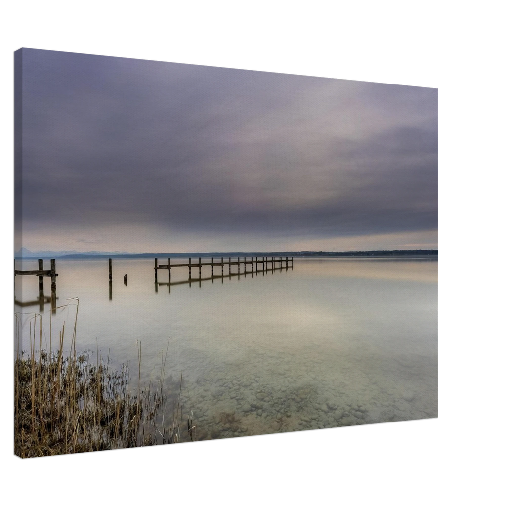 Leinwandbild mit ruhiger Ammersee-Landschaft in sanften Grau-, Beige- und Blautönen. Ein langer Holzsteg führt hinaus auf das spiegelglatte Wasser, während sich der weite Himmel mit seinen zarten Wolkenschichten im See widerspiegelt. Im Vordergrund sind Schilf und der klare, steinige Uferbereich sichtbar. Die hochwertige Leinwand auf 2 cm Keilrahmen bringt die feinen Farbverläufe und die stille Atmosphäre dieses zeitlosen Naturmotivs besonders harmonisch zur Geltung.