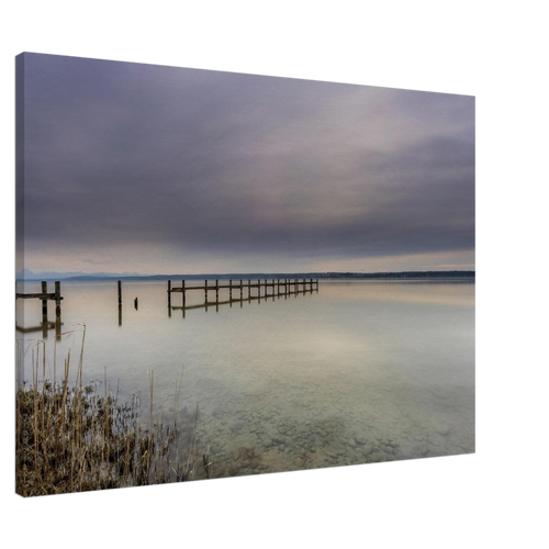 Leinwandbild mit ruhiger Ammersee-Landschaft in sanften Grau-, Beige- und Blautönen. Ein langer Holzsteg führt hinaus auf das spiegelglatte Wasser, während sich der weite Himmel mit seinen zarten Wolkenschichten im See widerspiegelt. Im Vordergrund sind Schilf und der klare, steinige Uferbereich sichtbar. Die hochwertige Leinwand auf 2 cm Keilrahmen bringt die feinen Farbverläufe und die stille Atmosphäre dieses zeitlosen Naturmotivs besonders harmonisch zur Geltung.