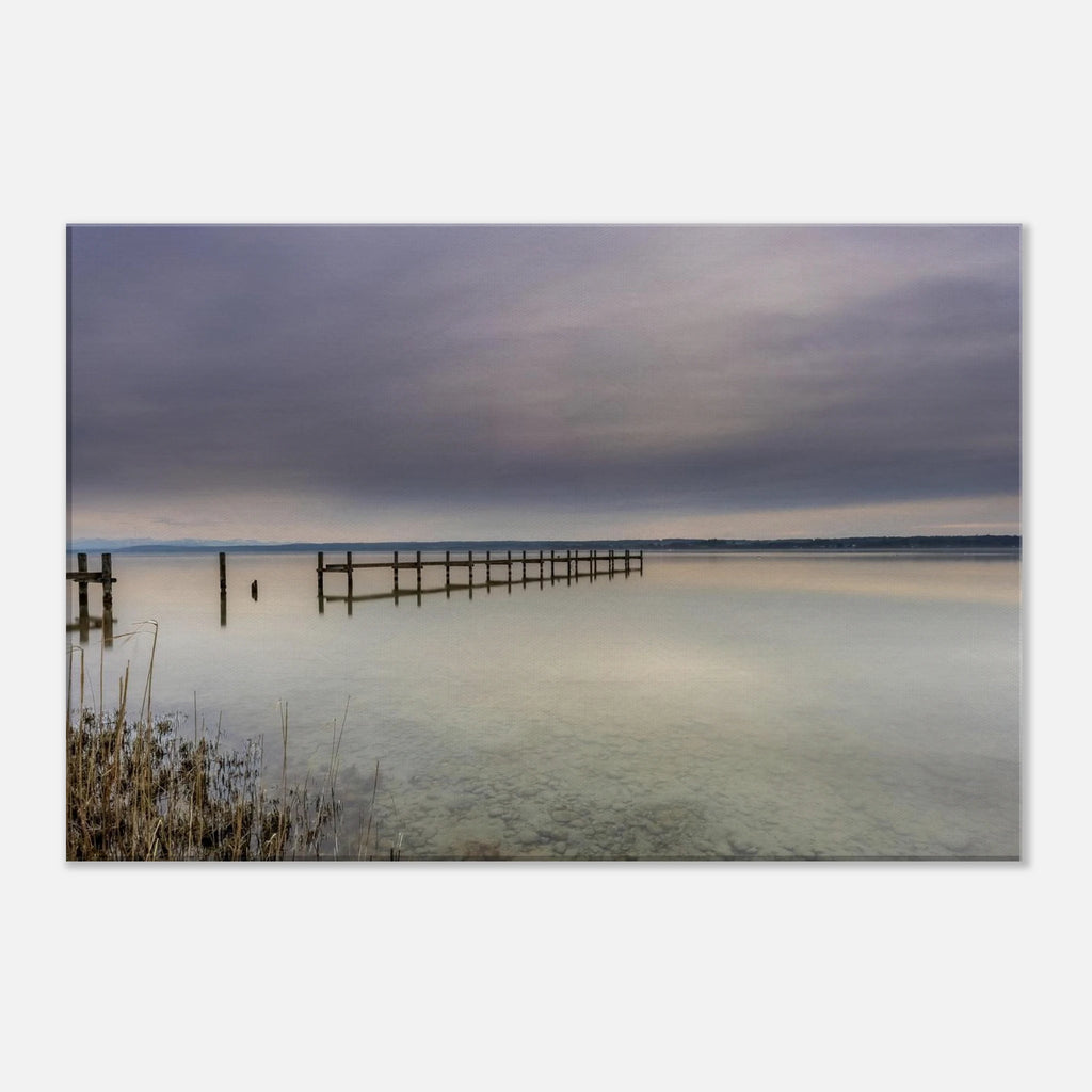 Leinwandbild mit ruhiger Ammersee-Landschaft in sanften Grau-, Beige- und Blautönen. Ein langer Holzsteg führt hinaus auf das spiegelglatte Wasser, während sich der weite Himmel mit seinen zarten Wolkenschichten im See widerspiegelt. Im Vordergrund sind Schilf und der klare, steinige Uferbereich sichtbar. Die hochwertige Leinwand auf 2 cm Keilrahmen bringt die feinen Farbverläufe und die stille Atmosphäre dieses zeitlosen Naturmotivs besonders harmonisch zur Geltung.