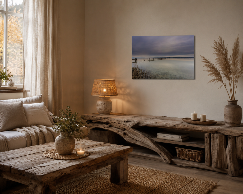 Gemütliche Wohnecke im Naturstil mit Möbeln aus Treibholz und warmen Erdtönen. Über einem rustikalen Sideboard hängt ein kleines, ungerahmtes Poster des Ammersees im Format 30 × 45 cm. Das Motiv zeigt einen stillen See mit verwittertem Steg unter einem weiten, wolkenverhangenen Himmel. Eine Tischlampe mit natürlichem Lampenschirm, Kerzen, Pampasgras und weiche Textilien schaffen eine ruhige, harmonische Atmosphäre voller Wärme und Gelassenheit.