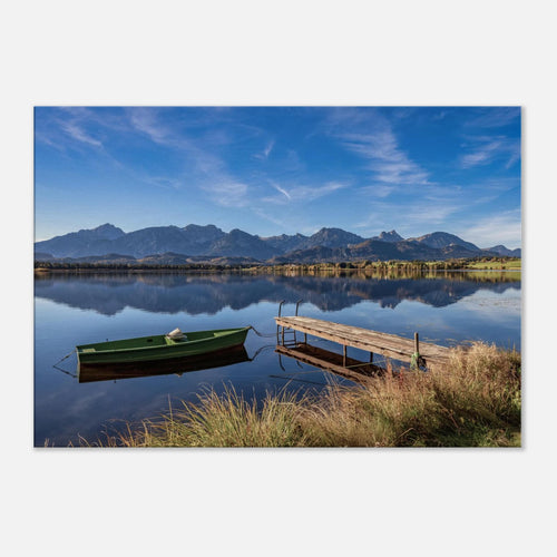 Leinwandbild auf 2 cm starkem Keilrahmen mit einer ruhigen Landschaftsaufnahme vom Hopfensee im Allgäu. Das Motiv zeigt ein grünes Ruderboot neben einem hölzernen Steg auf spiegelglattem Wasser. Im Hintergrund erheben sich die Berge unter einem klaren blauen Himmel mit feinen Wolkenstrukturen. Die seitliche Ansicht verdeutlicht die hochwertige Ausführung der Leinwand und die moderne Galerieoptik des Keilrahmens.
