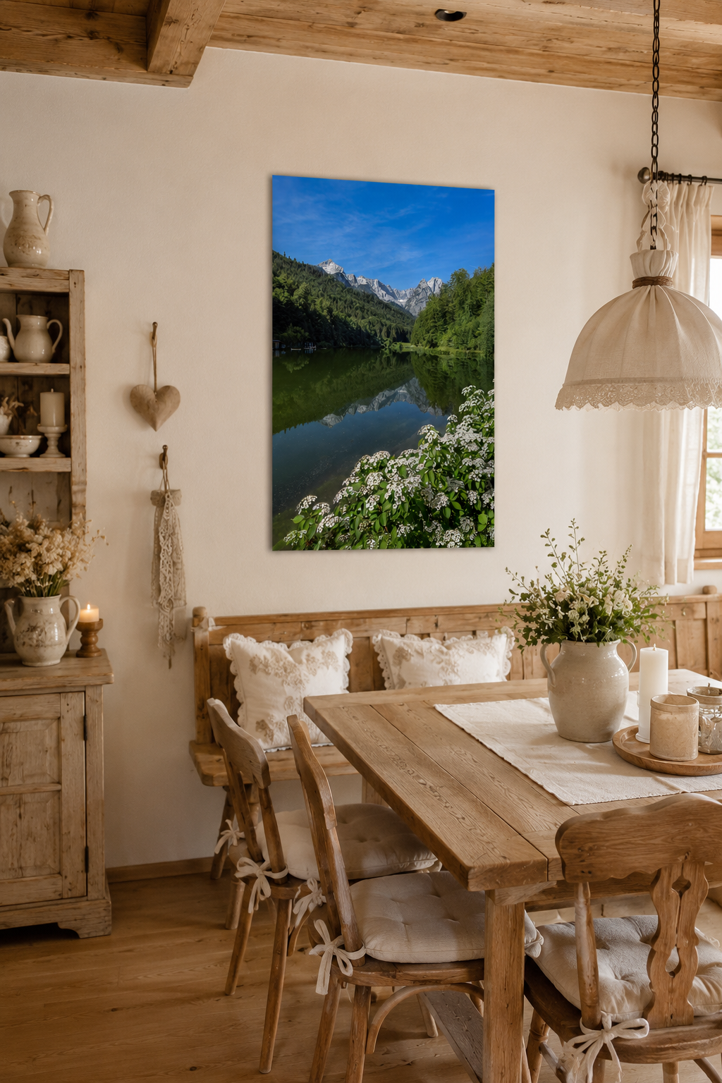 Helles Esszimmer im bayerischen Landhausstil mit Holzboden und Möbeln aus altem, naturbelassenem Holz. Cremefarbene Dekoration, weiße Spitzenkissen und natürliche Stoffe schaffen eine warme, einladende Atmosphäre. An der hellen Wand hängt ein ungerahmtes Poster im Hochformat 60 x 90 cm mit einer sommerlichen Ansicht des Riessersees bei Garmisch-Partenkirchen. Die Spiegelung der Berggipfel im ruhigen Wasser sowie die blühenden weißen Uferpflanzen fügen sich harmonisch in die natürlichen Farben des Raumes ein