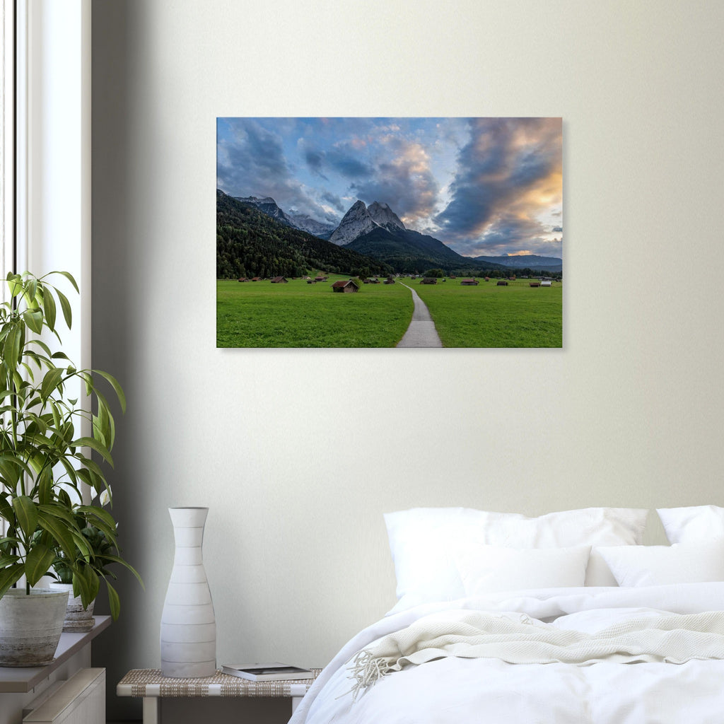 Landschaftsfotografie aus Garmisch-Partenkirchen mit Blick auf das imposante Wettersteingebirge. Ein schmaler Weg führt durch weitläufige grüne Wiesen mit traditionellen Heustadeln. Dramatische Wolken und sanftes Abendlicht tauchen die bayerische Alpenlandschaft in eine stimmungsvolle Atmosphäre.