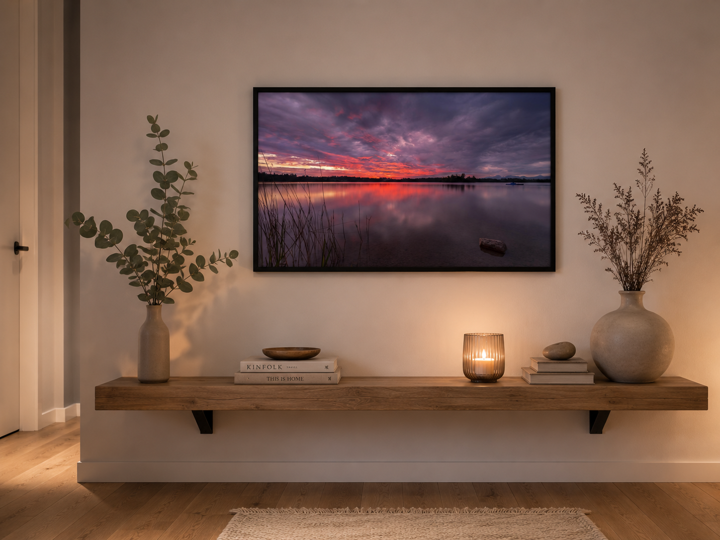 Großes Staffelsee-Poster im Format 70 × 100 cm im Querformat mit schwarzem Holzrahmen über einer massiven Balkenkonsole aus Naturholz. Das Motiv zeigt einen dramatischen Sonnenuntergang mit violett-rosa Wolkenformationen über dem ruhigen See und deren Spiegelung im Wasser. Auf der Konsole stehen dezente Naturdekoration, Kerzen und Vasen in harmonischen Beige- und Naturtönen. Heller Holzboden, warme Lichtstimmung und die auf das Wandbild abgestimmten Farben schaffen ein modernes, ruhiges und wohnliches Ambie