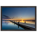Horizontales Wandbild mit schwarzem Holzrahmen. Das Motiv zeigt einen langen Holzsteg, der über das spiegelglatte Wasser eines Sees bis zum Horizont führt. Die untergehende Sonne taucht Himmel und Wasser in warme Gold-, Orange- und Blautöne. Durch die Langzeitbelichtung wirkt die Wasseroberfläche besonders ruhig und glatt. Der schwarze Holzrahmen setzt einen modernen, eleganten Akzent und betont die klare Linienführung des Motivs.
