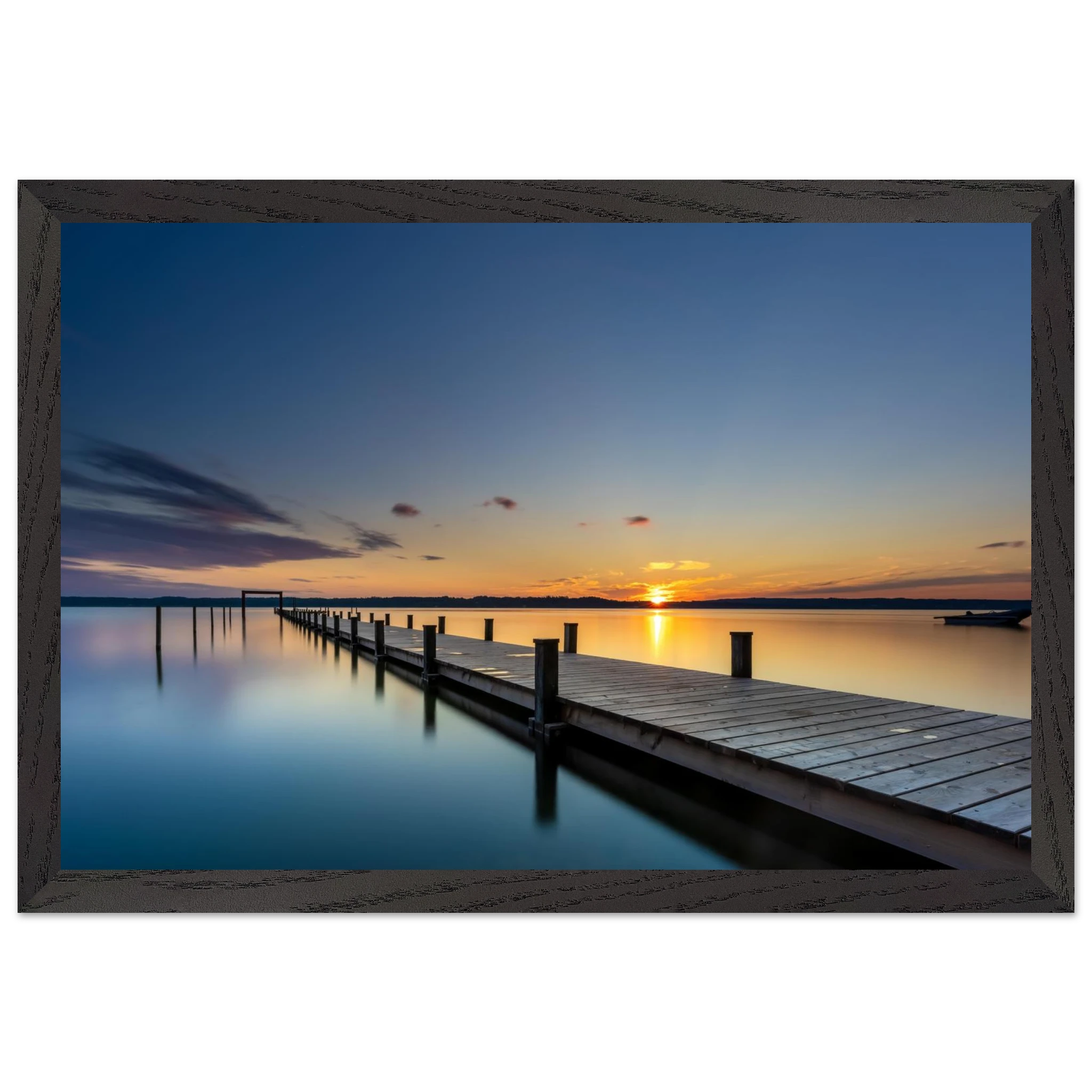 Horizontales Wandbild mit schwarzem Holzrahmen. Das Motiv zeigt einen langen Holzsteg, der über das spiegelglatte Wasser eines Sees bis zum Horizont führt. Die untergehende Sonne taucht Himmel und Wasser in warme Gold-, Orange- und Blautöne. Durch die Langzeitbelichtung wirkt die Wasseroberfläche besonders ruhig und glatt. Der schwarze Holzrahmen setzt einen modernen, eleganten Akzent und betont die klare Linienführung des Motivs.