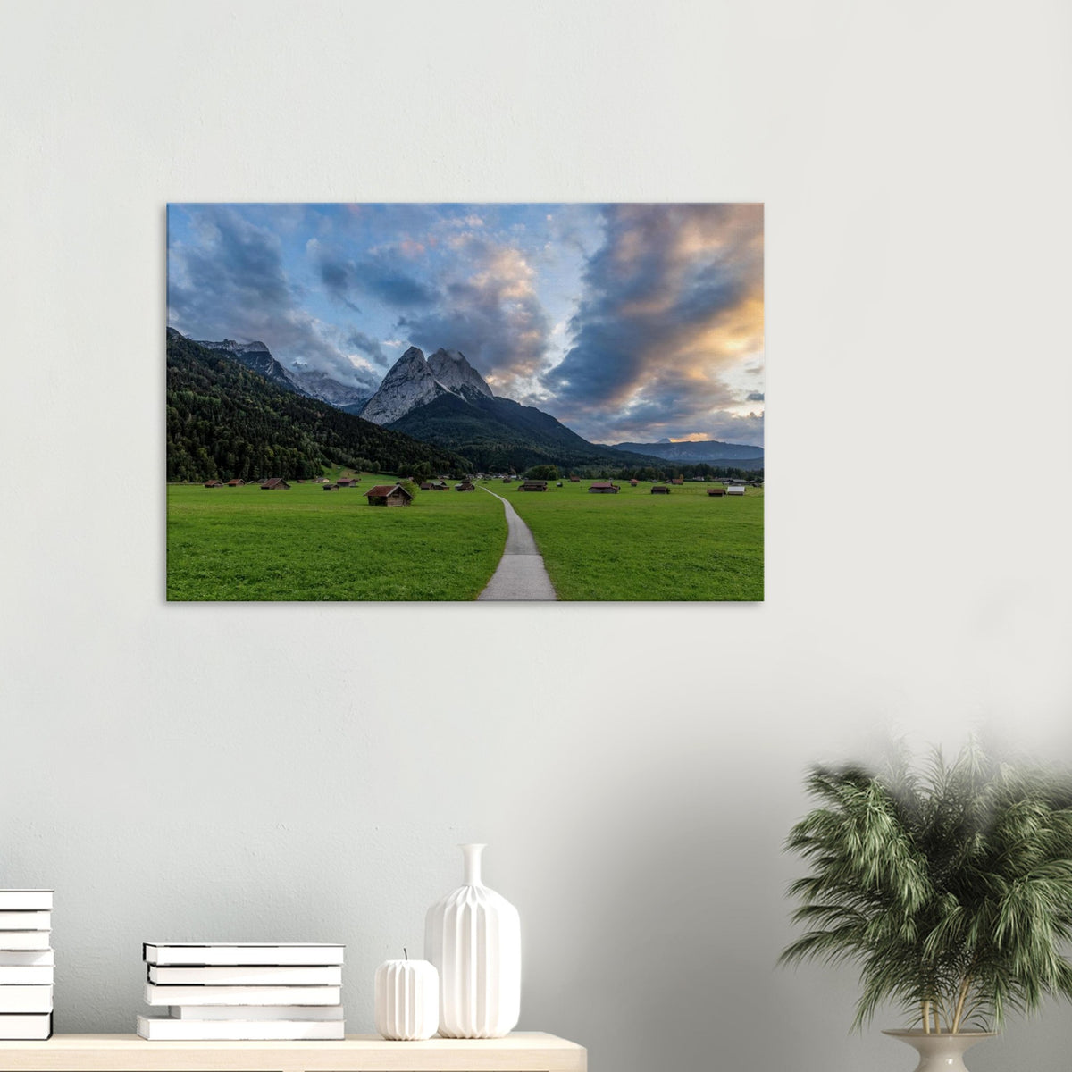Landschaftsfotografie aus Garmisch-Partenkirchen mit Blick auf das imposante Wettersteingebirge. Ein schmaler Weg führt durch weitläufige grüne Wiesen mit traditionellen Heustadeln. Dramatische Wolken und sanftes Abendlicht tauchen die bayerische Alpenlandschaft in eine stimmungsvolle Atmosphäre.
