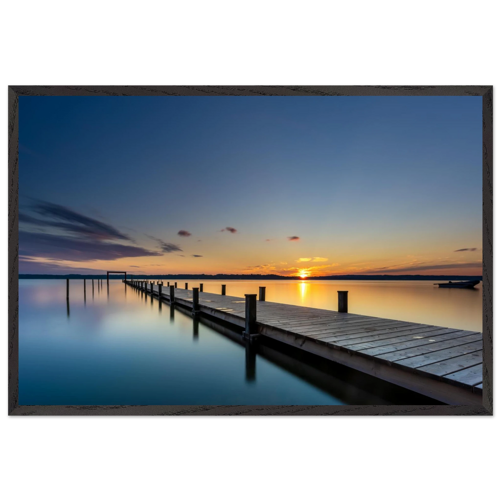 Horizontales Wandbild mit schwarzem Holzrahmen. Das Motiv zeigt einen langen Holzsteg, der über das spiegelglatte Wasser eines Sees bis zum Horizont führt. Die untergehende Sonne taucht Himmel und Wasser in warme Gold-, Orange- und Blautöne. Durch die Langzeitbelichtung wirkt die Wasseroberfläche besonders ruhig und glatt. Der schwarze Holzrahmen setzt einen modernen, eleganten Akzent und betont die klare Linienführung des Motivs.