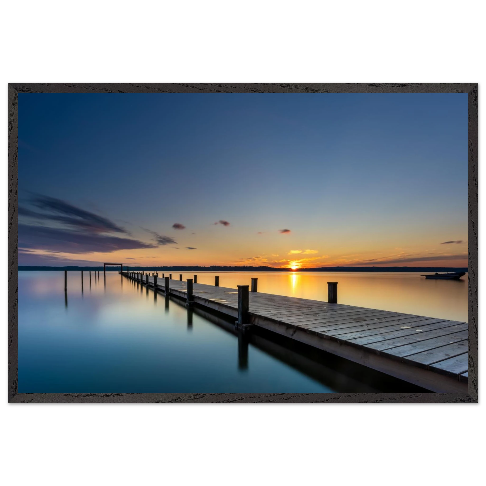 Horizontales Wandbild mit schwarzem Holzrahmen. Das Motiv zeigt einen langen Holzsteg, der über das spiegelglatte Wasser eines Sees bis zum Horizont führt. Die untergehende Sonne taucht Himmel und Wasser in warme Gold-, Orange- und Blautöne. Durch die Langzeitbelichtung wirkt die Wasseroberfläche besonders ruhig und glatt. Der schwarze Holzrahmen setzt einen modernen, eleganten Akzent und betont die klare Linienführung des Motivs.
