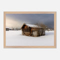 Horizontales Poster mit naturfarbenem Holzrahmen. Das Motiv zeigt eine alte, verwitterte Holzhütte auf einer verschneiten Winterwiese. Im Hintergrund sind weitere Gebäude und ein Kirchturm zu sehen, während dunkle Wolken über der winterlichen Landschaft liegen. Ein heller Lichtstrahl durchbricht die Wolkendecke und verleiht der Szene eine ruhige, stimmungsvolle Atmosphäre. Der natürliche Holzrahmen unterstreicht den rustikalen Charakter des Wintermotivs.