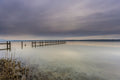 Ruhige Stimmung am Ammersee mit einem langen Holzsteg, der weit in das spiegelglatte Wasser hinausführt. Im Vordergrund sind Schilf und die klar sichtbaren Steine des flachen Uferbereichs zu erkennen. Über dem See liegt ein sanft bewölkter Himmel in zarten Grau-, Blau- und Beigetönen, dessen Licht sich im Wasser spiegelt. Die weite, reduzierte Landschaft vermittelt Stille, Gelassenheit und die zeitlose Schönheit eines friedlichen Moments am Ammersee.