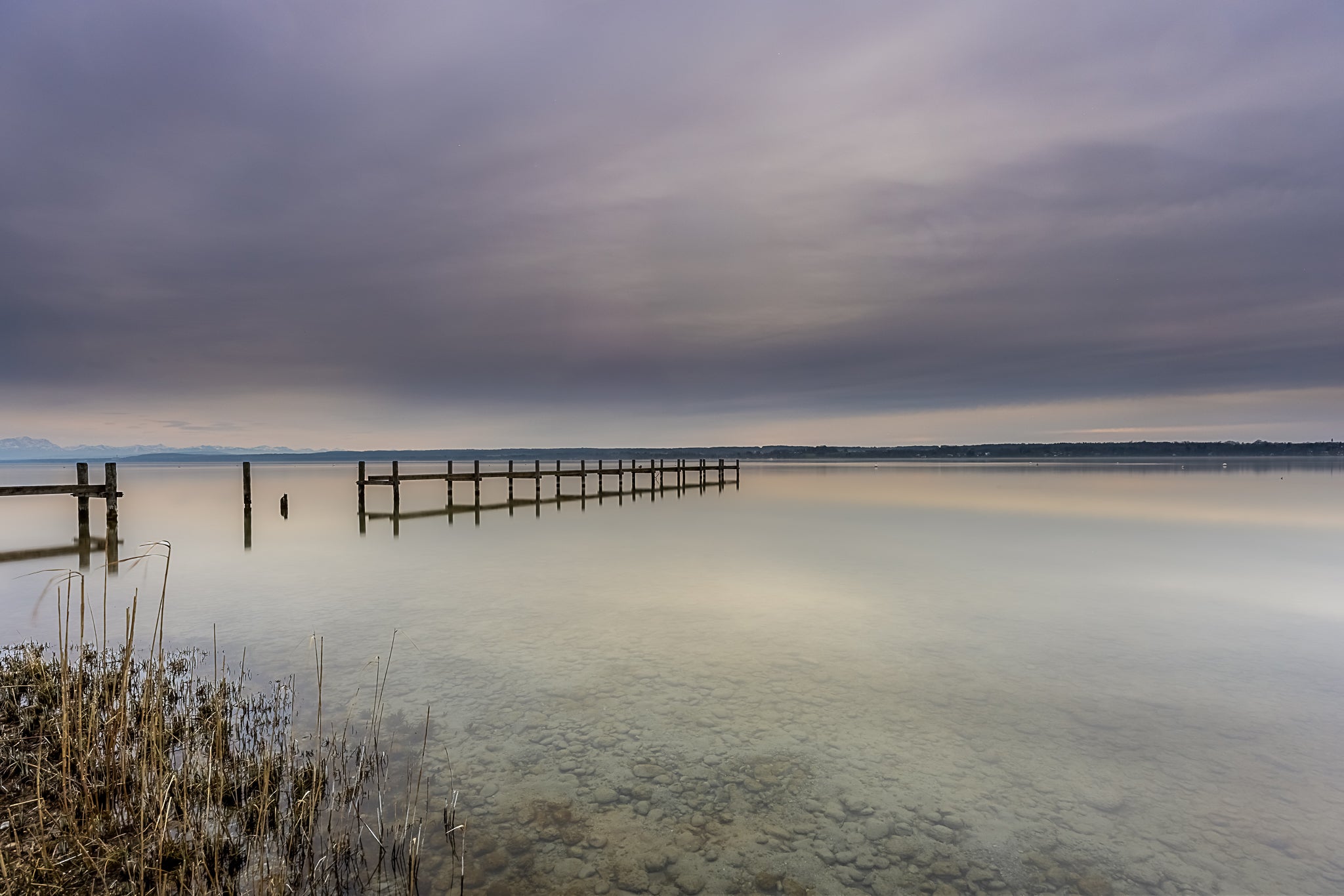 Ruhige Stimmung am Ammersee mit einem langen Holzsteg, der weit in das spiegelglatte Wasser hinausführt. Im Vordergrund sind Schilf und die klar sichtbaren Steine des flachen Uferbereichs zu erkennen. Über dem See liegt ein sanft bewölkter Himmel in zarten Grau-, Blau- und Beigetönen, dessen Licht sich im Wasser spiegelt. Die weite, reduzierte Landschaft vermittelt Stille, Gelassenheit und die zeitlose Schönheit eines friedlichen Moments am Ammersee.