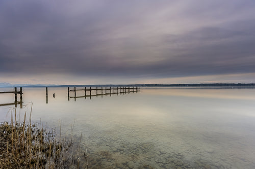 Ruhige Stimmung am Ammersee mit einem langen Holzsteg, der weit in das spiegelglatte Wasser hinausführt. Im Vordergrund sind Schilf und die klar sichtbaren Steine des flachen Uferbereichs zu erkennen. Über dem See liegt ein sanft bewölkter Himmel in zarten Grau-, Blau- und Beigetönen, dessen Licht sich im Wasser spiegelt. Die weite, reduzierte Landschaft vermittelt Stille, Gelassenheit und die zeitlose Schönheit eines friedlichen Moments am Ammersee.