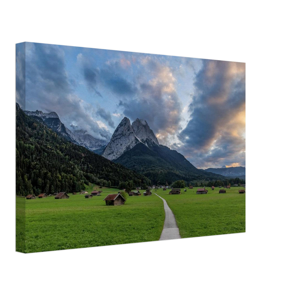 Landschaftsfotografie aus Garmisch-Partenkirchen mit Blick auf das imposante Wettersteingebirge. Ein schmaler Weg führt durch weitläufige grüne Wiesen mit traditionellen Heustadeln. Dramatische Wolken und sanftes Abendlicht tauchen die bayerische Alpenlandschaft in eine stimmungsvolle Atmosphäre.