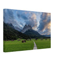 Landschaftsfotografie aus Garmisch-Partenkirchen mit Blick auf das imposante Wettersteingebirge. Ein schmaler Weg führt durch weitläufige grüne Wiesen mit traditionellen Heustadeln. Dramatische Wolken und sanftes Abendlicht tauchen die bayerische Alpenlandschaft in eine stimmungsvolle Atmosphäre.