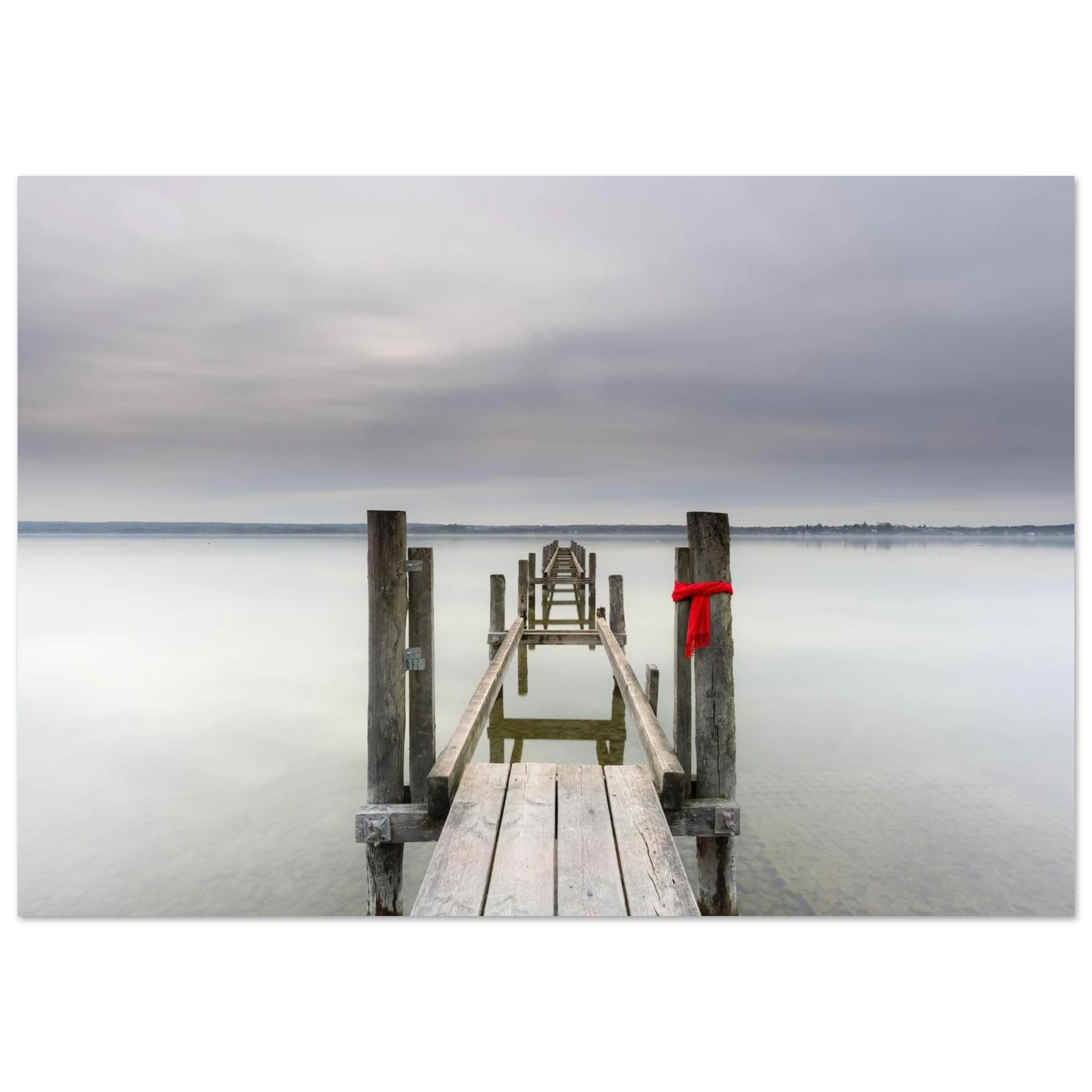Alter Holzsteg am Ammersee mit einem leuchtend roten Schal, der an einem Pfosten befestigt ist. Die ruhige Langzeitbelichtung, das glatte Wasser und der sanft bewölkte Himmel schaffen eine stille, minimalistische Atmosphäre in Oberbayern.
