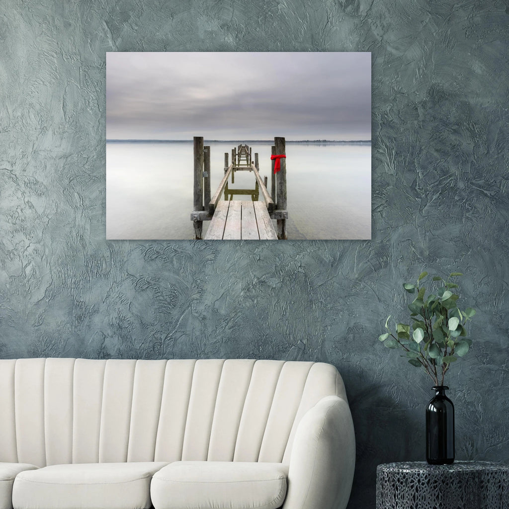 Alter Holzsteg am Ammersee mit einem leuchtend roten Schal, der an einem Pfosten befestigt ist. Die ruhige Langzeitbelichtung, das glatte Wasser und der sanft bewölkte Himmel schaffen eine stille, minimalistische Atmosphäre in Oberbayern.