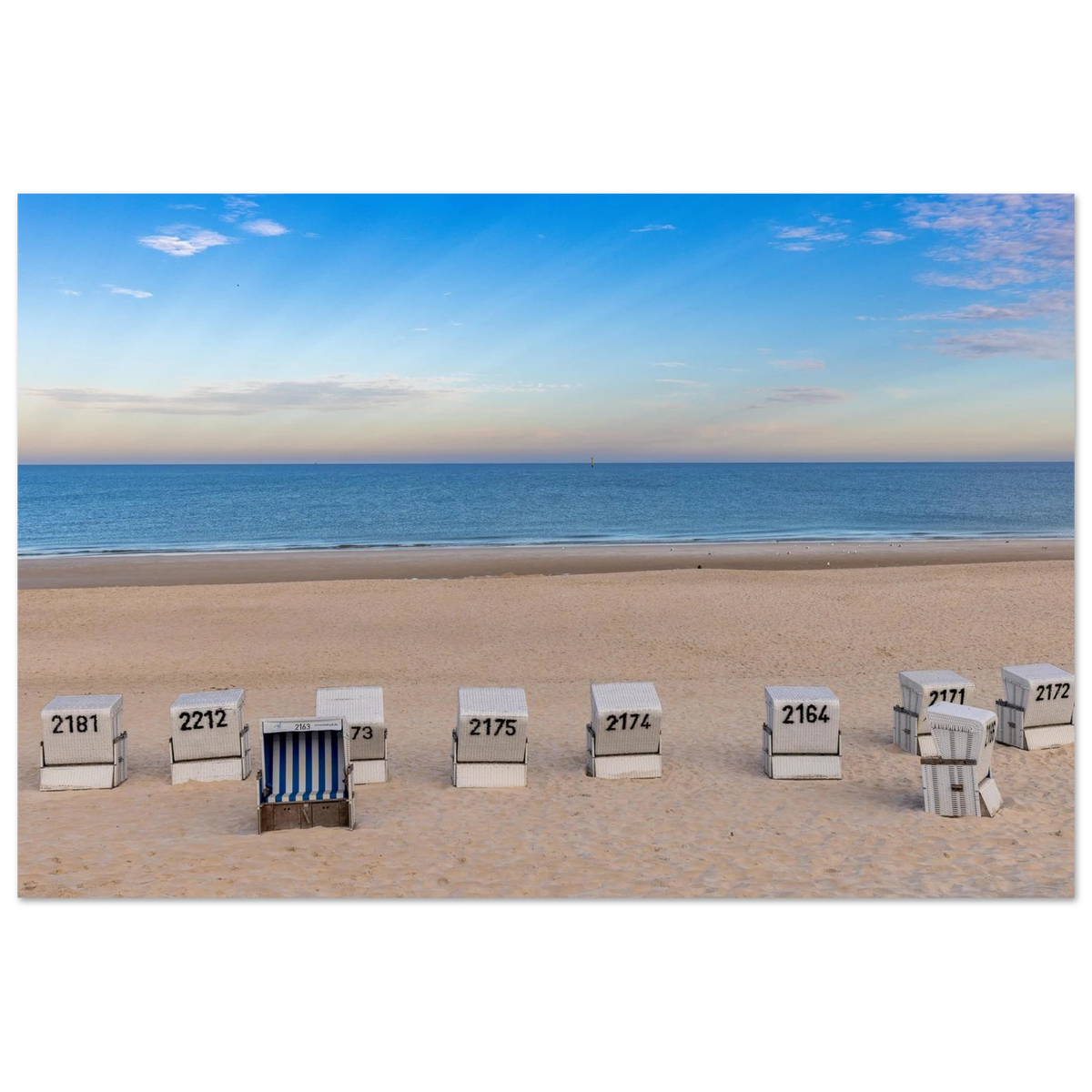 Das Motiv zeigt einen weiten, ruhigen Strand in Westerland auf Sylt in einer klaren, sommerlichen Stimmung. Im Vordergrund liegt der helle, fein strukturierte Sand, in dem mehrere weiße Strandkörbe in einer lockeren Reihe stehen. Einer davon ist geöffnet und mit blau-weiß gestreiftem Innenstoff ein besonderer Blickfang.
