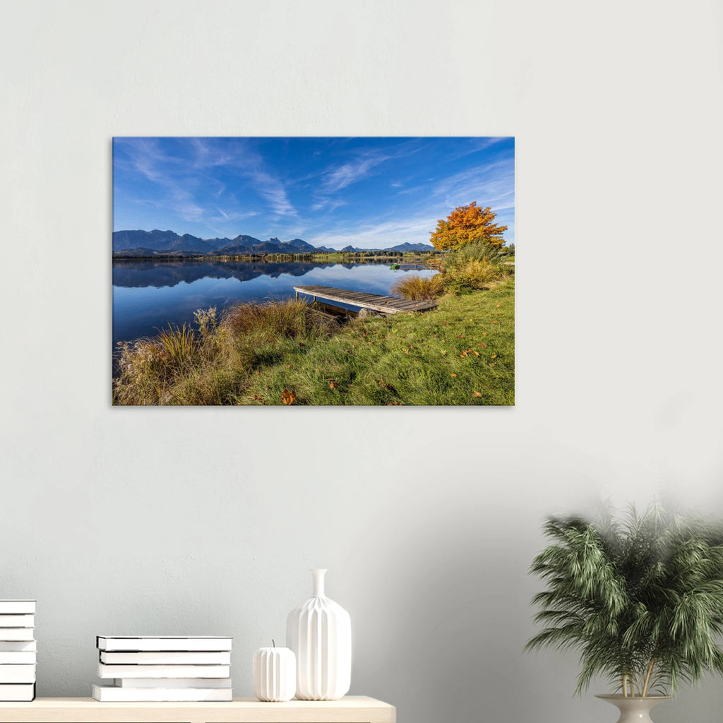 bEin goldener Herbsttag im Allgäu. Der Hopfensee liegt still vor der beeindruckenden Kulisse der Berge, das klare Wasser trägt ihre Spiegelungen und am Ufer leuchtet der Herbst in warmen Farben. Der kleine Holzsteg führt hinaus in diese besondere Ruhe, die man manchmal nur in der Natur findet.