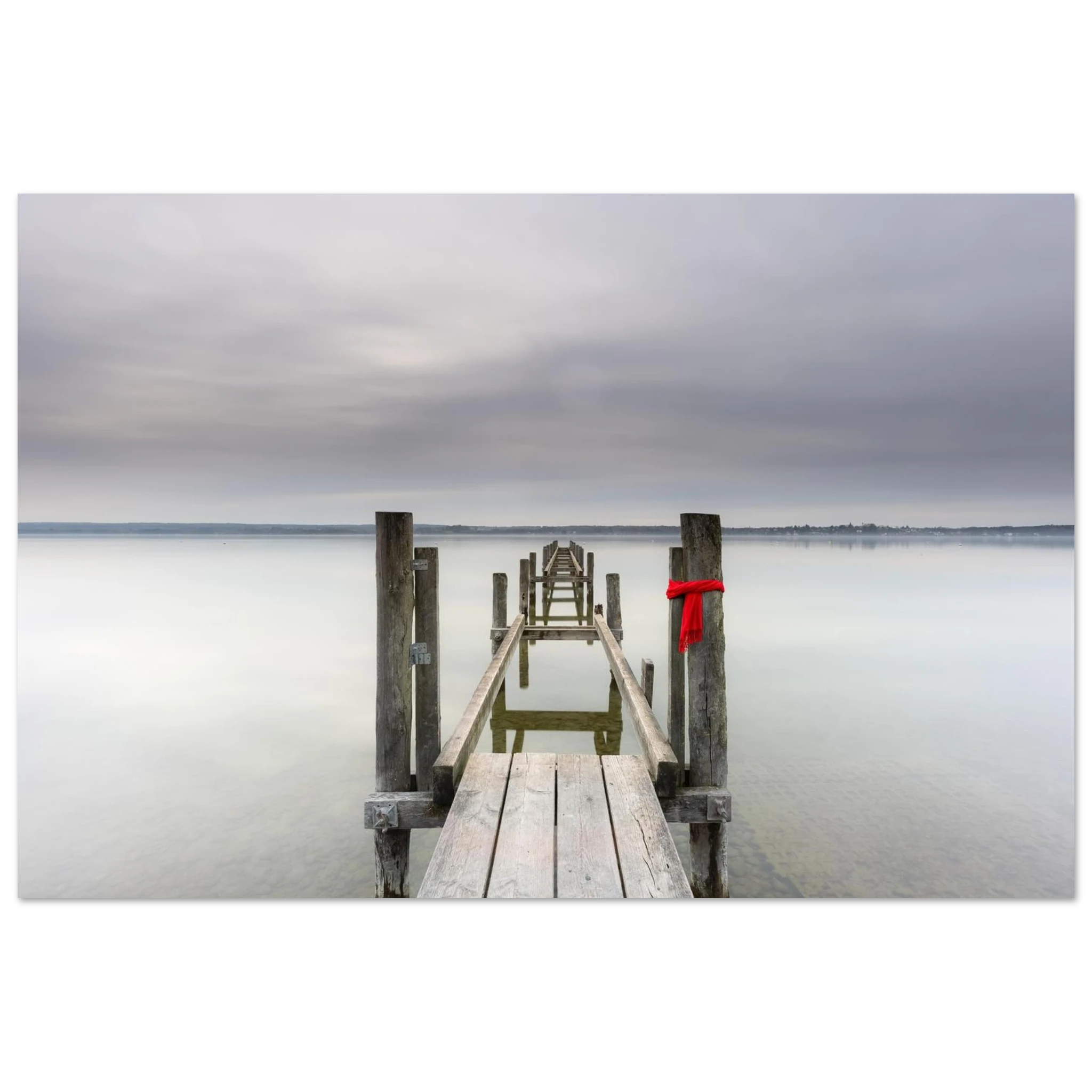 Alter Holzsteg am Ammersee mit einem leuchtend roten Schal, der an einem Pfosten befestigt ist. Die ruhige Langzeitbelichtung, das glatte Wasser und der sanft bewölkte Himmel schaffen eine stille, minimalistische Atmosphäre in Oberbayern.