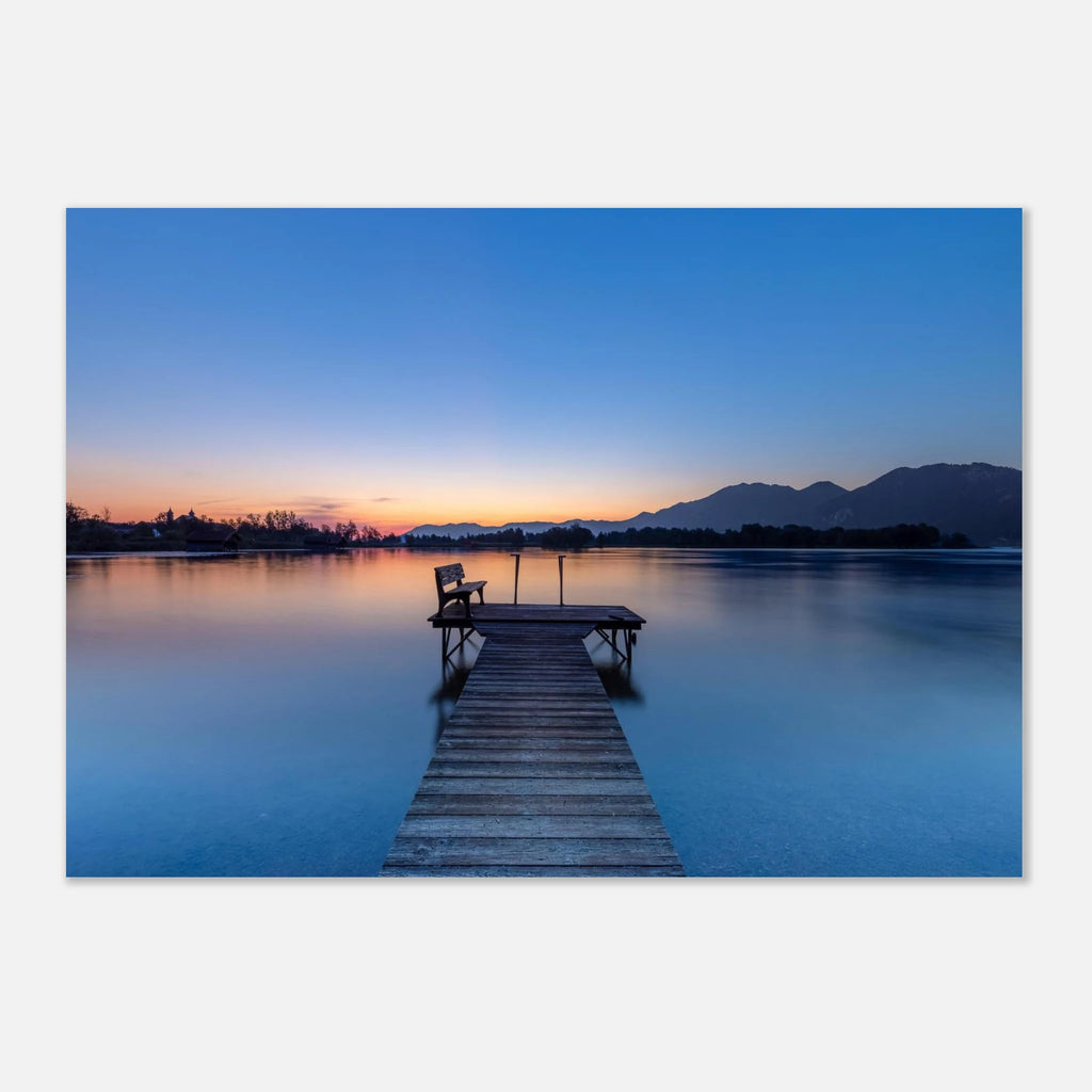 Stimmungsvoller Morgen am Kochelsee in Oberbayern. Ein hölzerner Steg mit einer kleinen Bank führt in das spiegelglatte Wasser. Sanfte Blau- und Orangetöne der Morgendämmerung spiegeln sich im See, während sich die Silhouetten der Berge am Horizont abzeichnen. Eine ruhige Landschaftsfotografie in der Blauen Stunde.