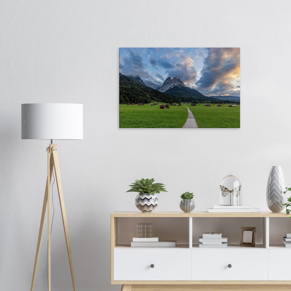 Landschaftsfotografie aus Garmisch-Partenkirchen mit Blick auf das imposante Wettersteingebirge. Ein schmaler Weg führt durch weitläufige grüne Wiesen mit traditionellen Heustadeln. Dramatische Wolken und sanftes Abendlicht tauchen die bayerische Alpenlandschaft in eine stimmungsvolle Atmosphäre.
